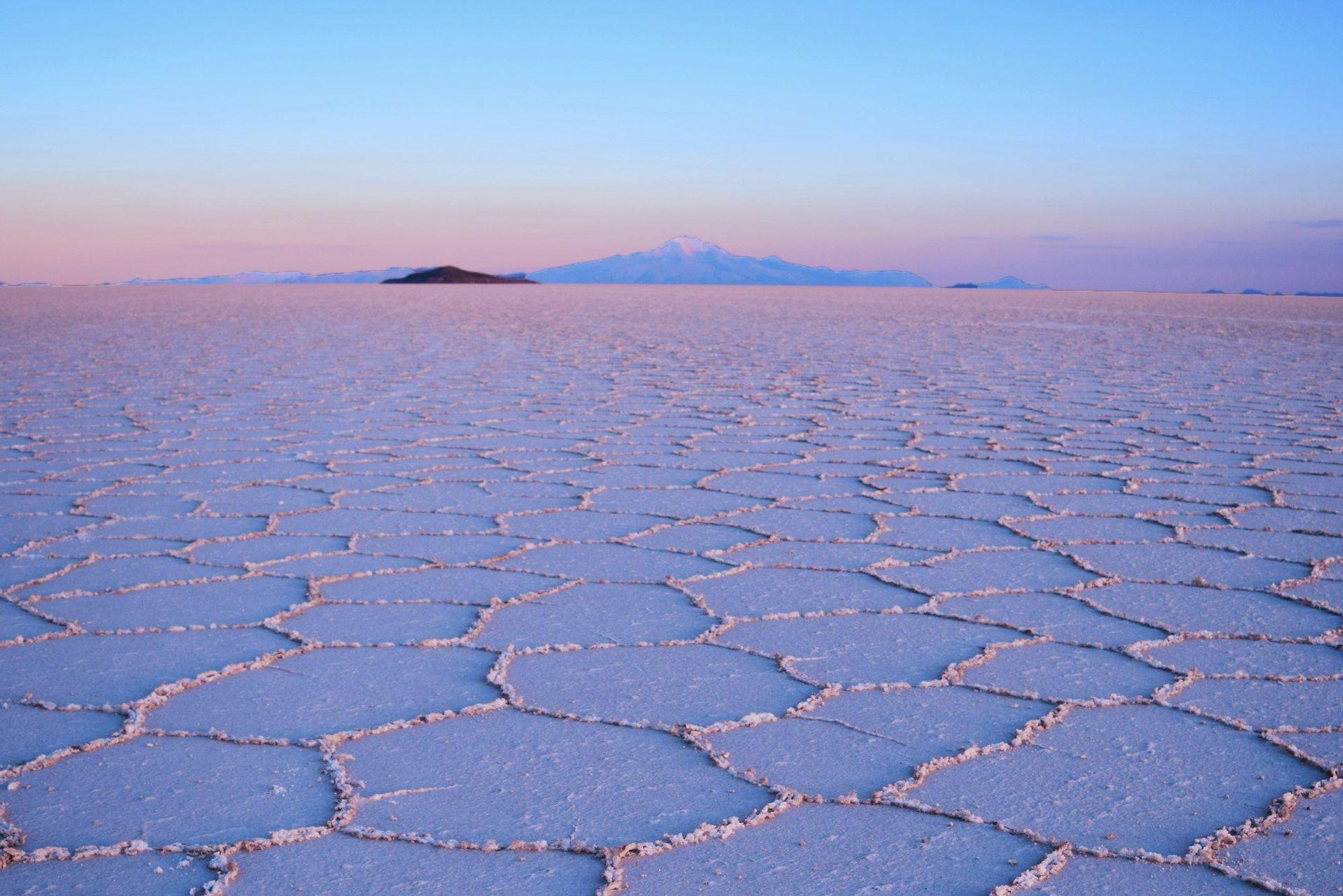 A vast salt flat with natural hexagonal patterns stretches towards distant mountains under a blue and pink sky at dusk.