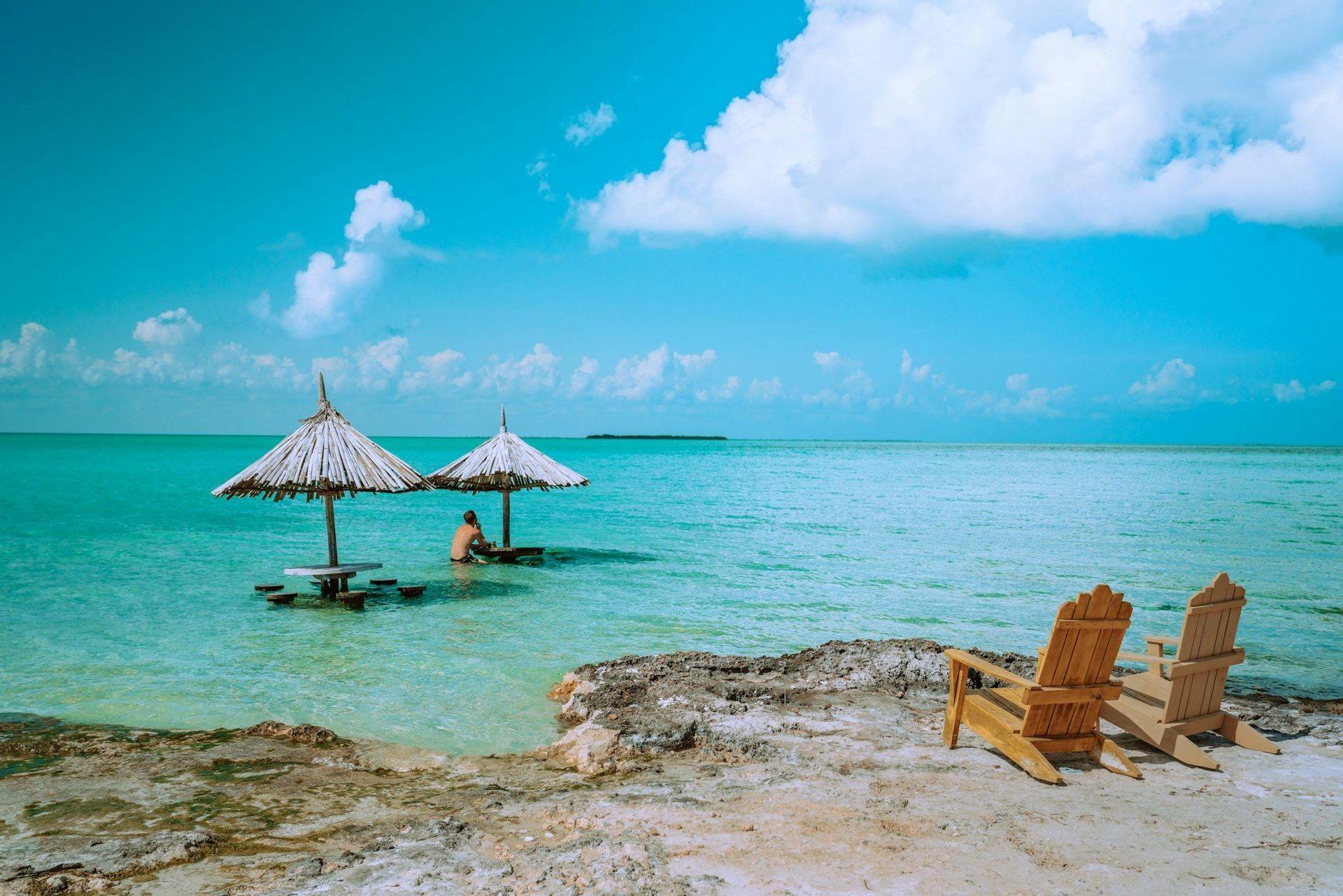 Two empty wooden chairs on a rocky shore overlook a person sitting at a table with a thatched umbrella in turquoise water.