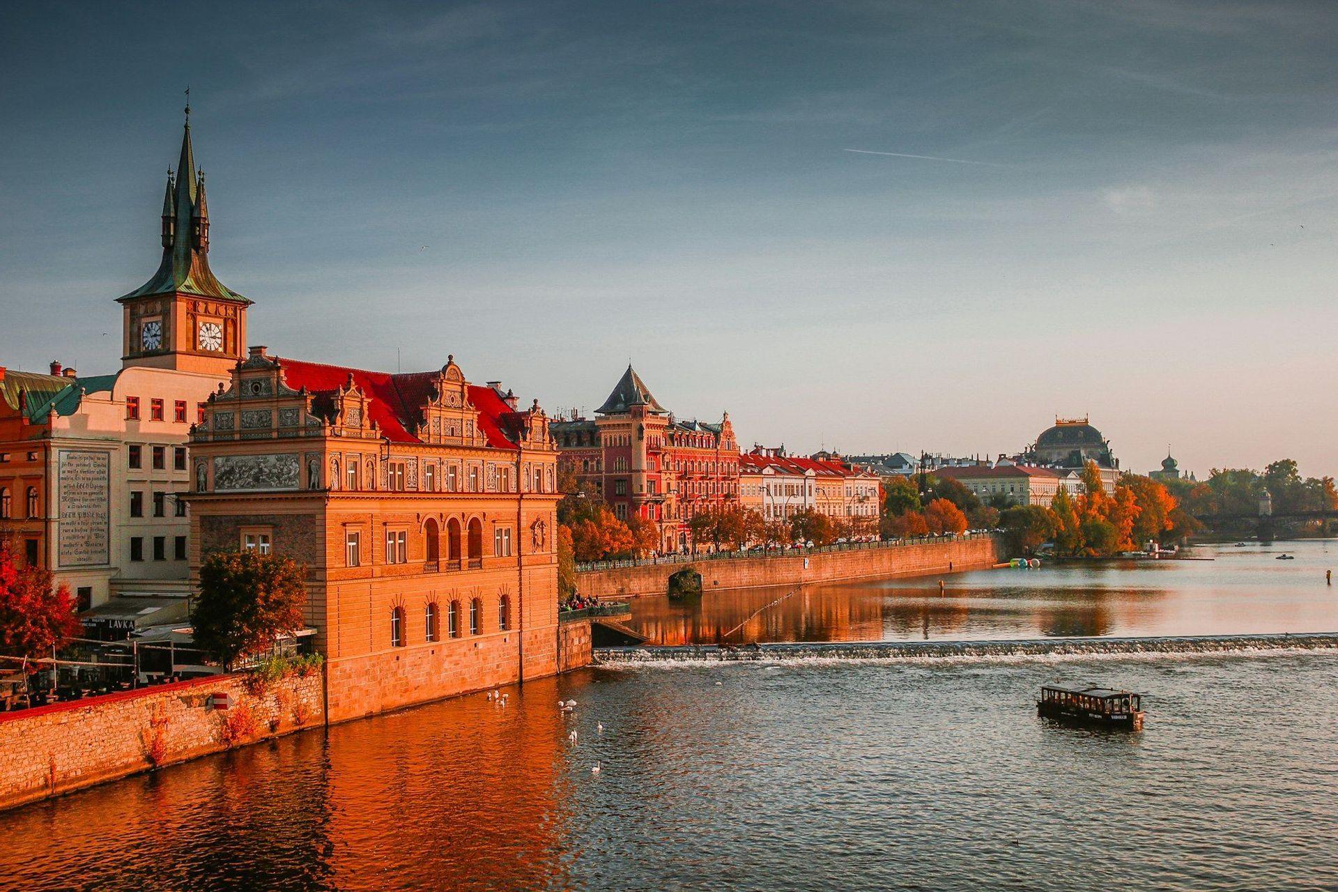 A cityscape of historic buildings with red roofs and a clock tower along a river at sunset, with a small boat on the water.