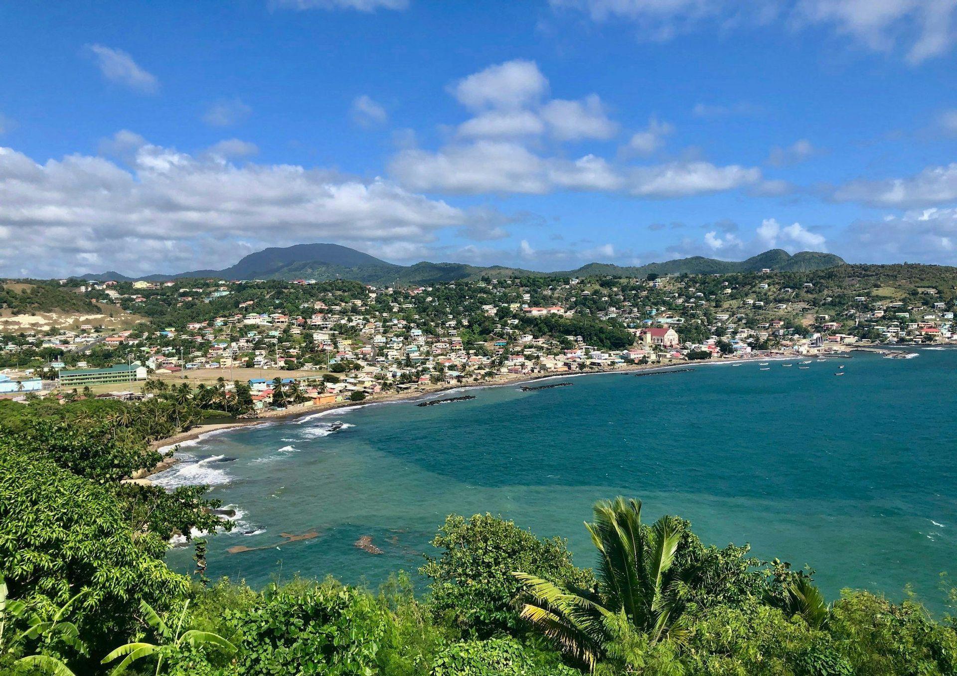 Vista aérea de un pueblo costero enclavado en una bahía, con colinas verdes que se alzan al fondo bajo un cielo parcialmente nublado.