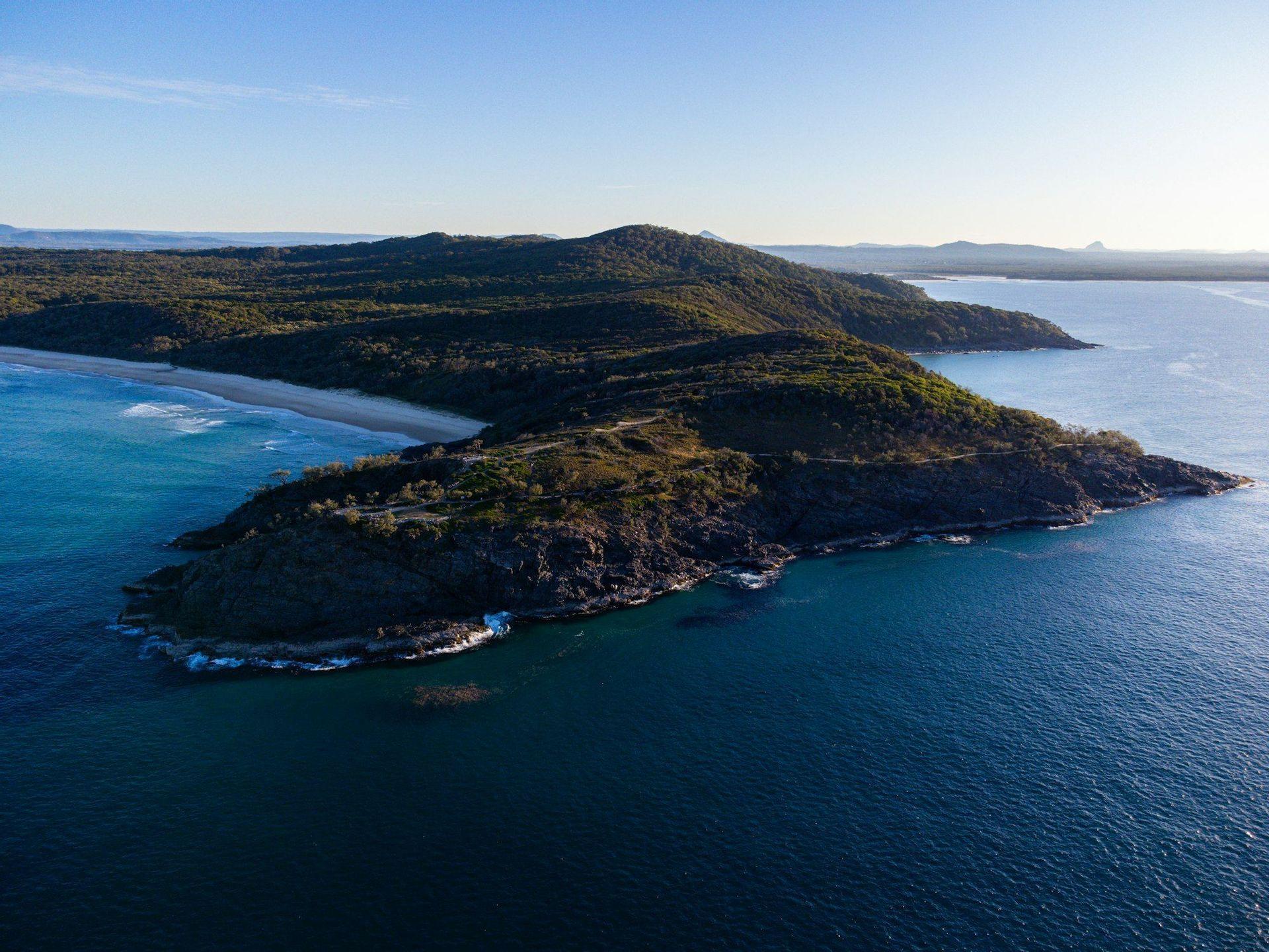 Una vista aérea de una península verde y soleada con una costa rocosa que se encuentra con un mar azul profundo, con una playa de arena a su izquierda.