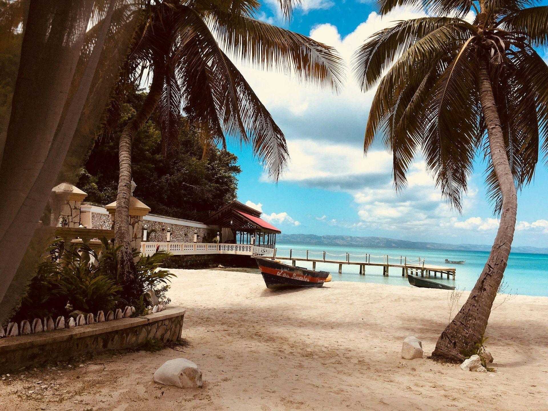 Una playa tropical de arena con palmeras, un barco en la orilla y un muelle de madera que se extiende sobre aguas turquesas bajo un cielo azul.