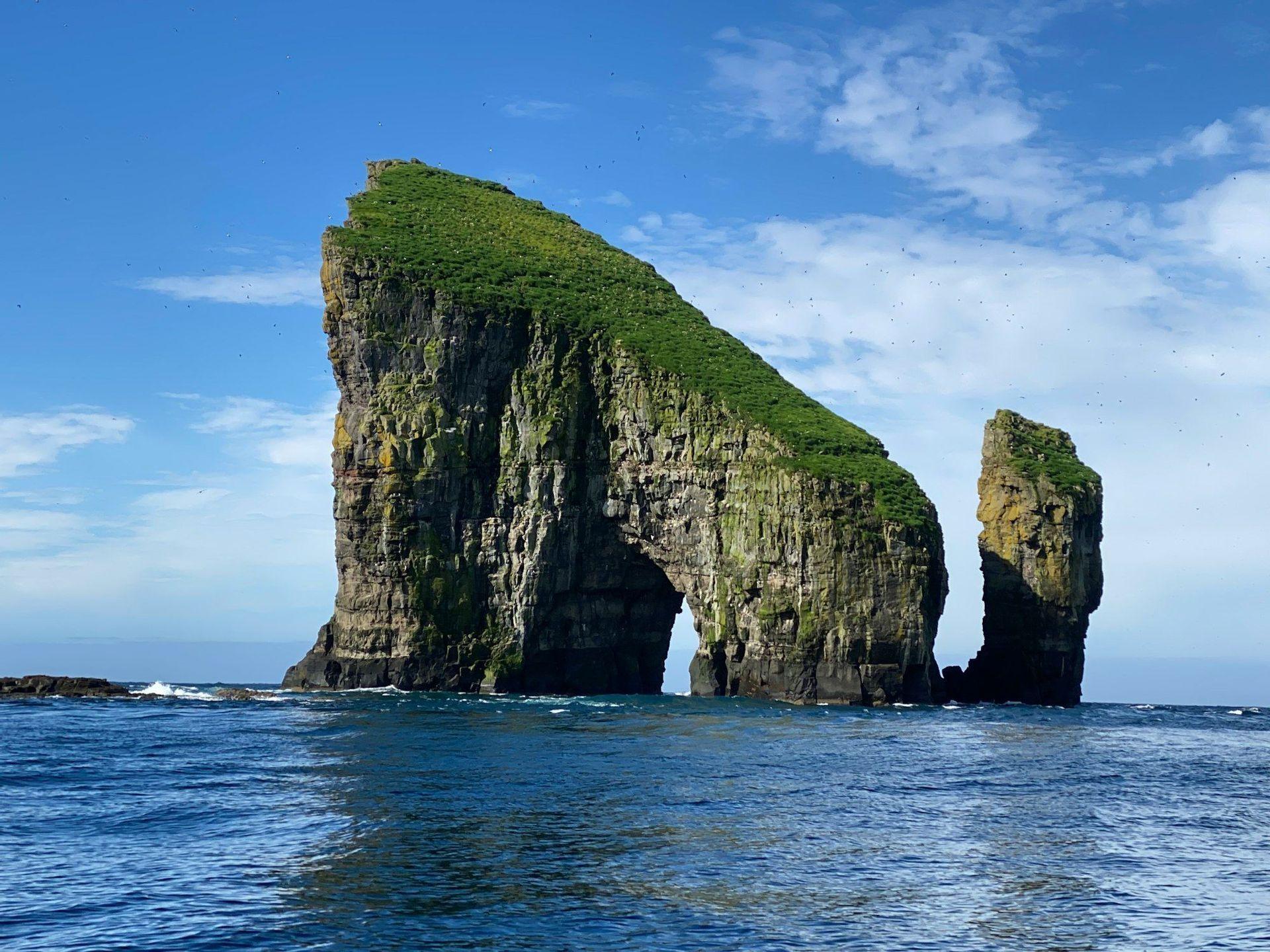 Un imponente farallón cubierto de hierba con un arco natural se eleva desde el océano azul bajo un cielo despejado con nubes dispersas.