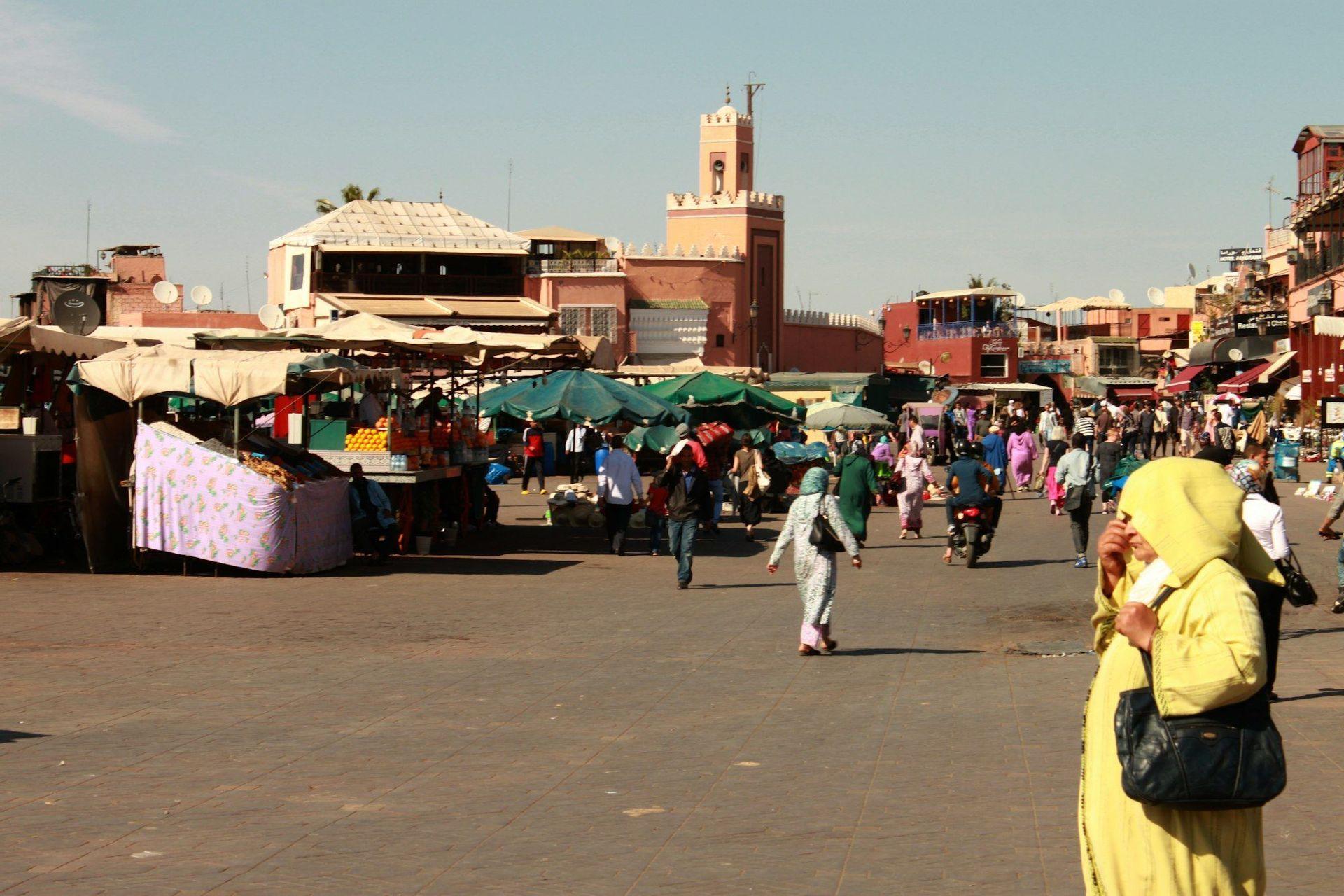 Una animada plaza de mercado al aire libre llena de gente paseando entre puestos bajo un cielo despejado y soleado, con edificios de terracota al fondo.