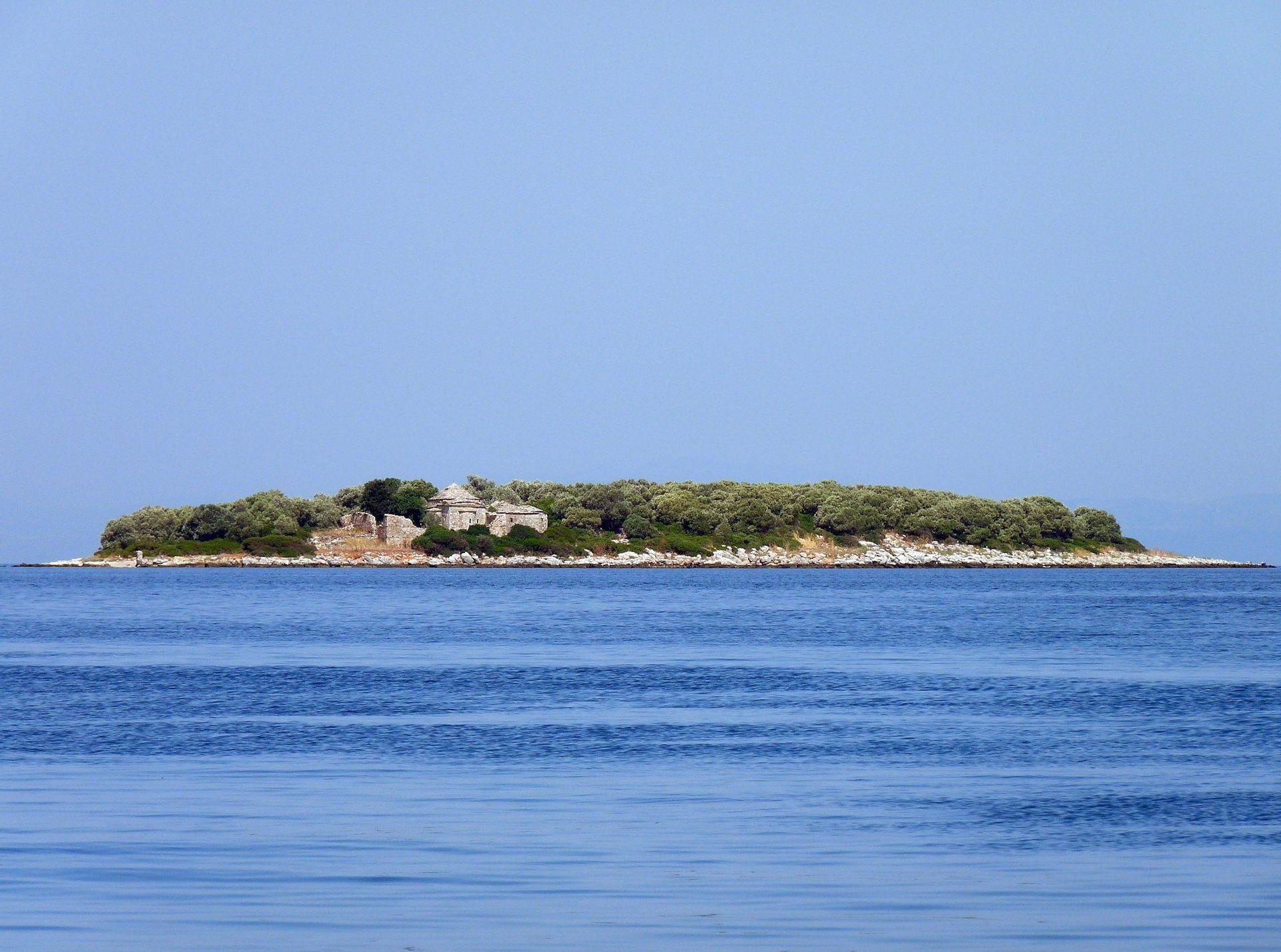 Eine kleine, bewaldete Insel mit Steinruinen liegt inmitten eines ruhigen blauen Meeres unter klarem Himmel.