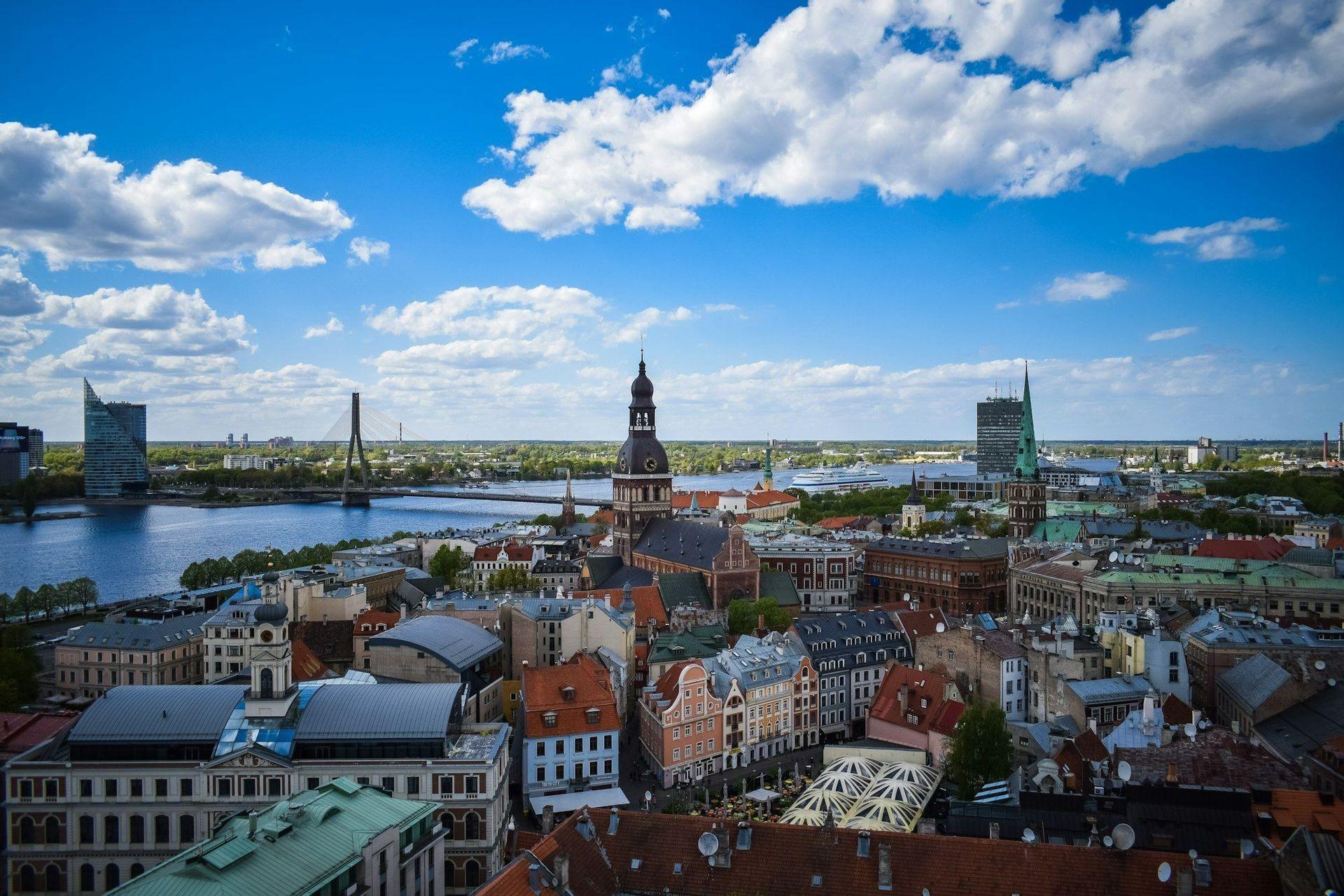 Eine Luftaufnahme einer historischen Stadt mit einem großen Kirchturm, einem breiten Fluss und einer modernen Brücke unter blauem Himmel mit Wolken.