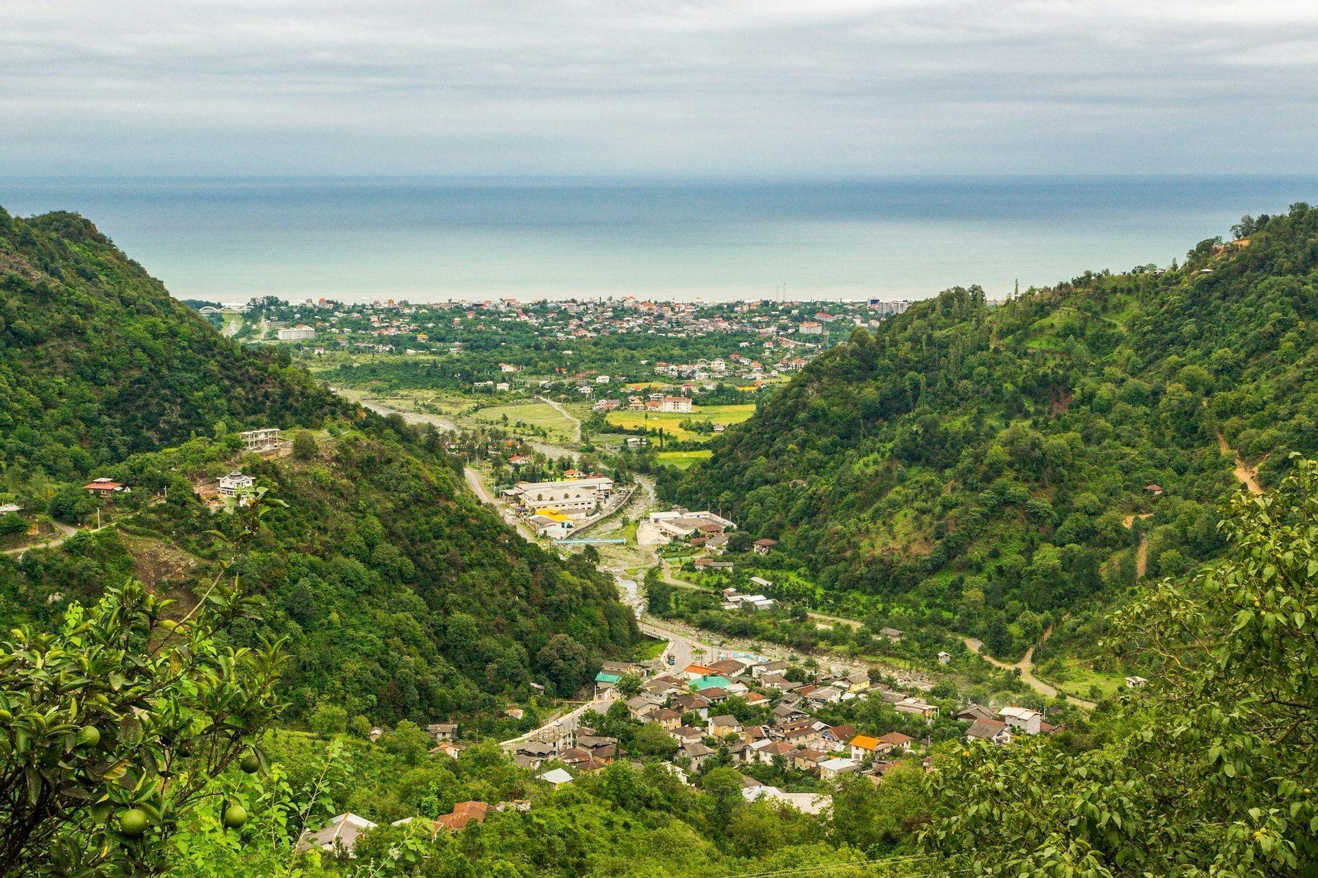 Luftaufnahme einer Stadt, eingebettet in einem üppig grünen Tal zwischen bewaldeten Hügeln, mit dem Meer im Hintergrund.