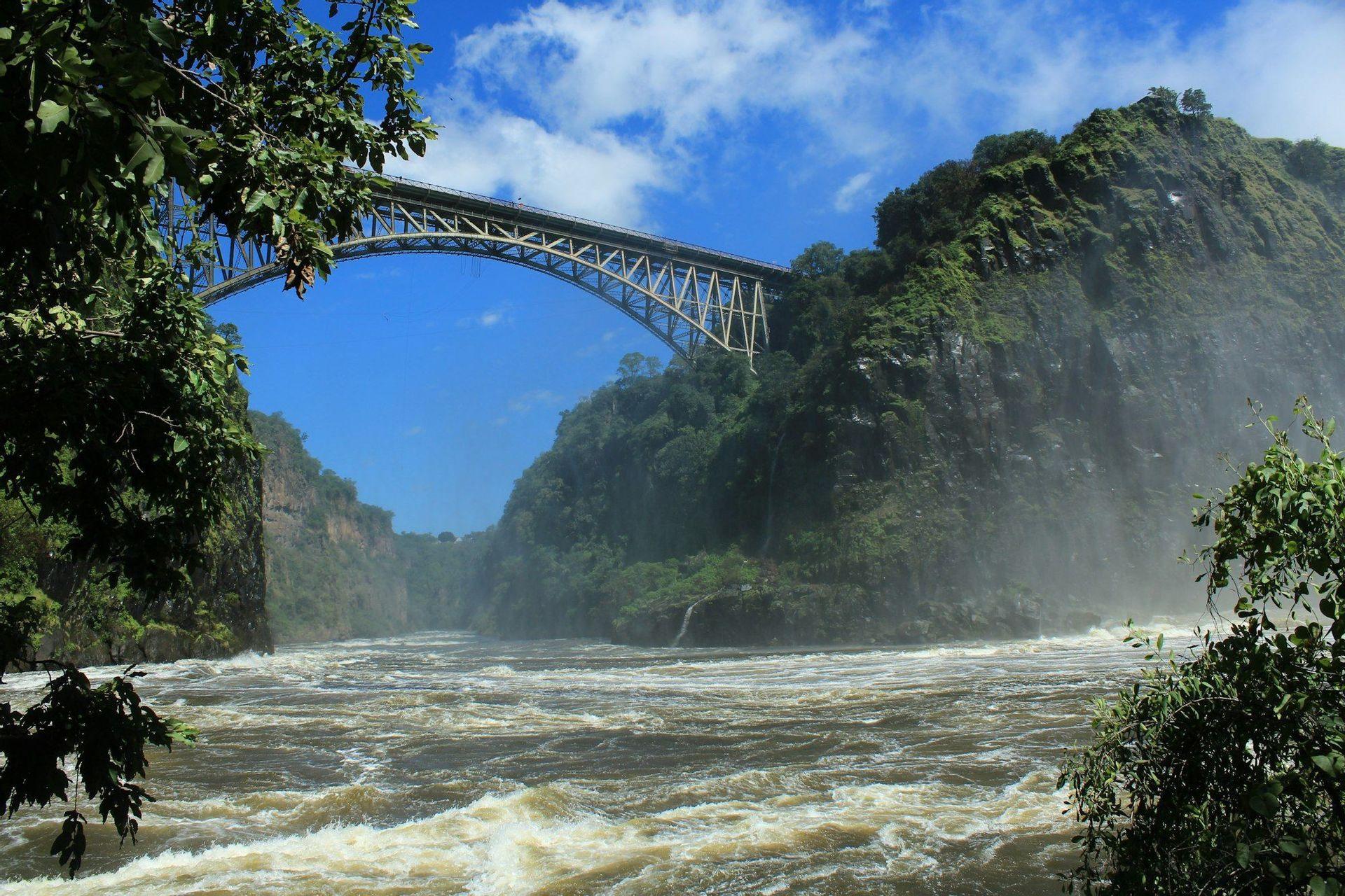 Eine Stahlbogenbrücke überspannt einen reißenden Fluss, der durch eine tiefe, grüne Schlucht unter einem teilweise bewölkten Himmel fließt.