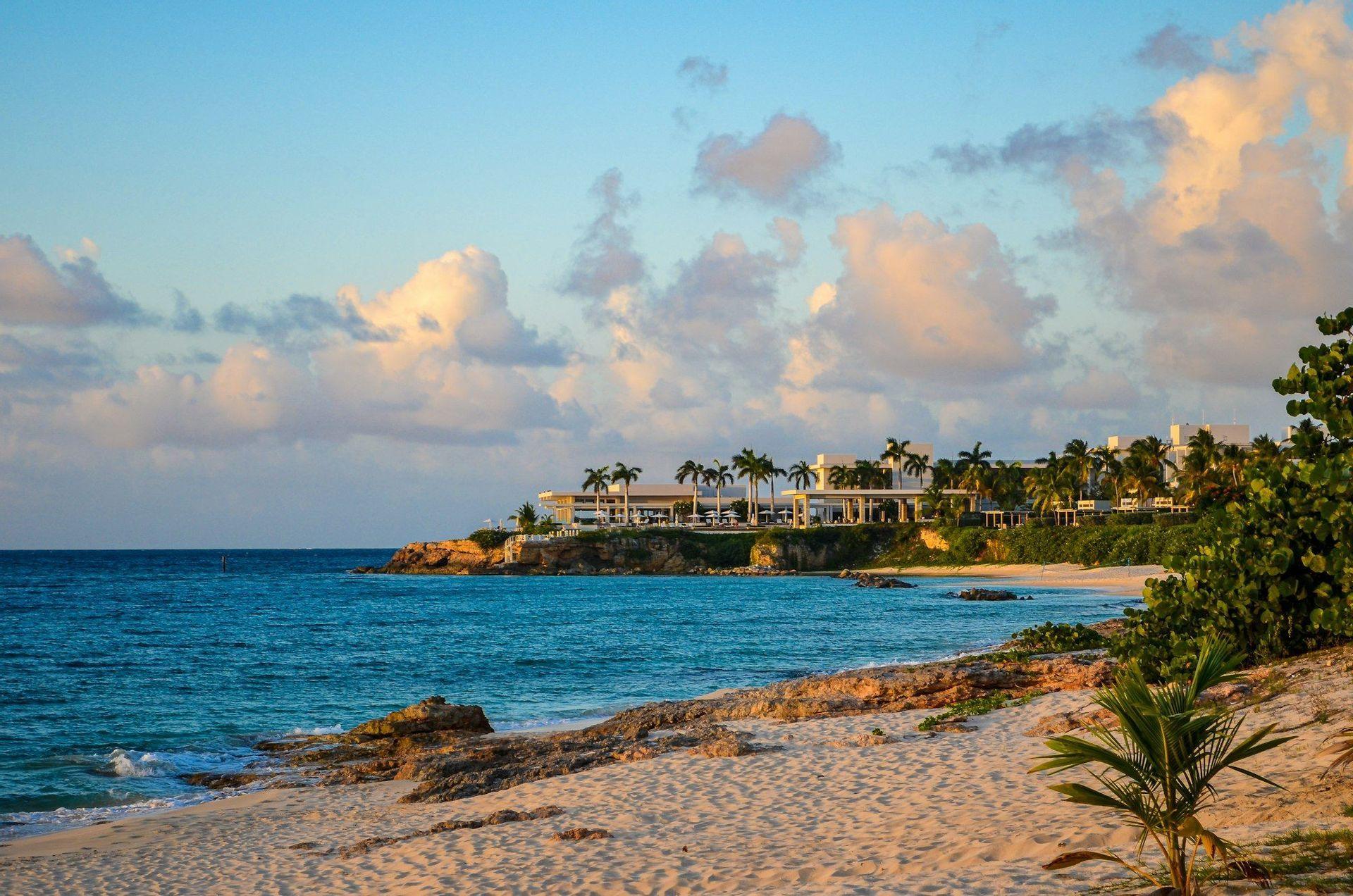 Un complexe hôtelier blanc avec des palmiers est perché sur un littoral rocheux, à côté d'une plage de sable et de l'océan turquoise, au coucher du soleil.