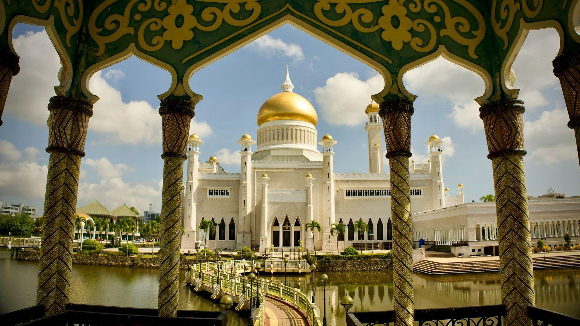 Une mosquée blanche au dôme doré proéminent est encadrée par des arches vertes et dorées ornementées, avec un plan d'eau au premier plan.