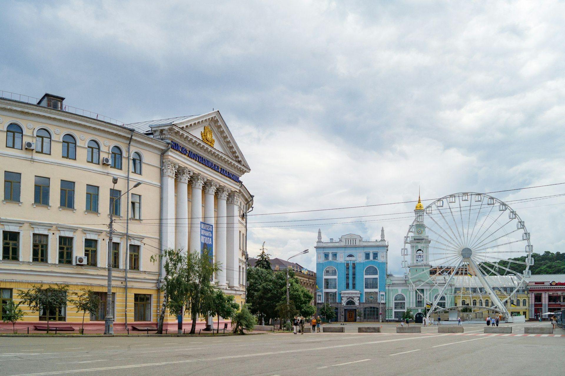 Une grande place de ville avec des bâtiments classiques colorés et une grande roue sous un ciel nuageux.