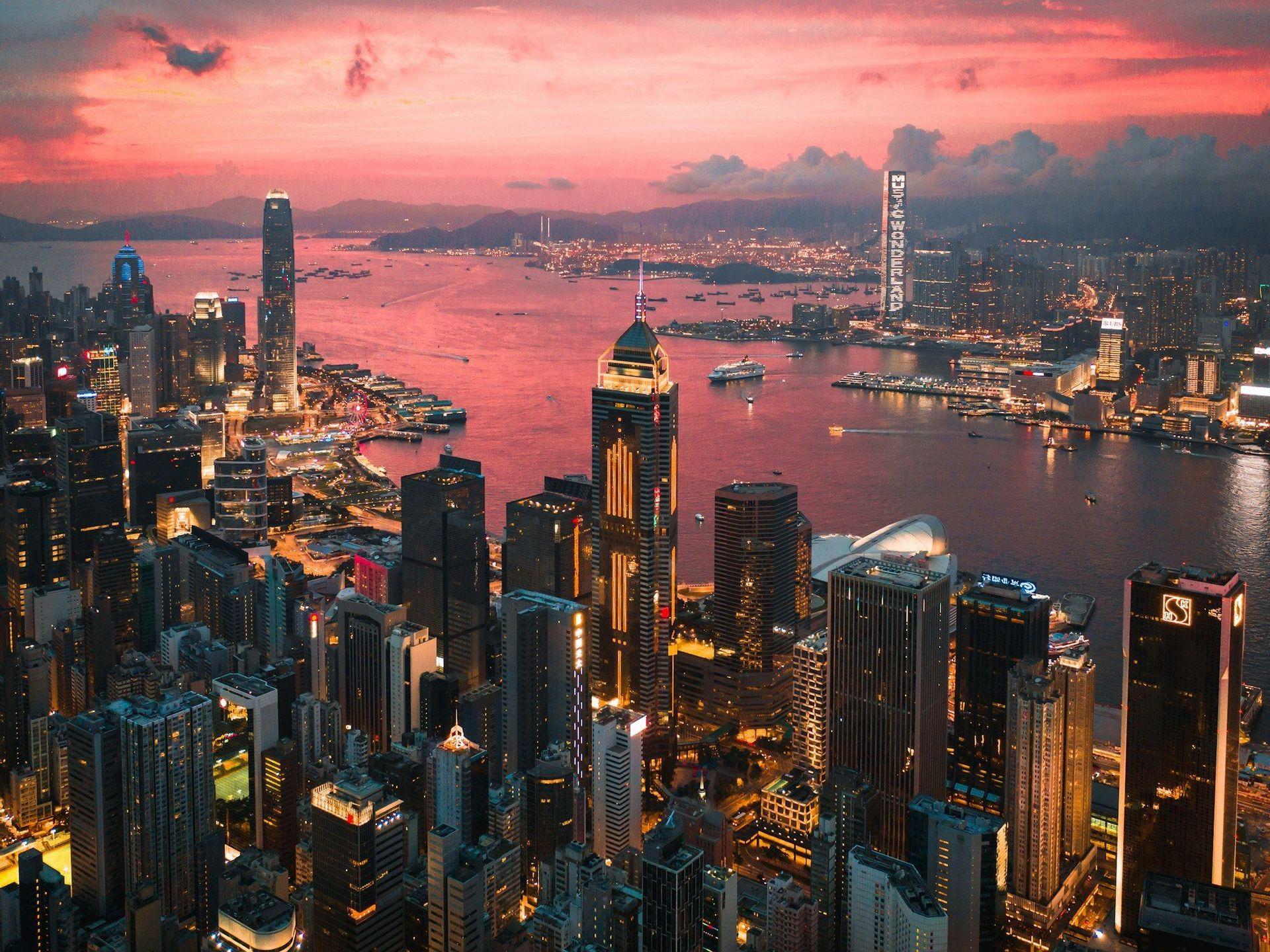 Vue aérienne d'une ville tentaculaire et illuminée, bordant un port sous un ciel de coucher de soleil rose éclatant.