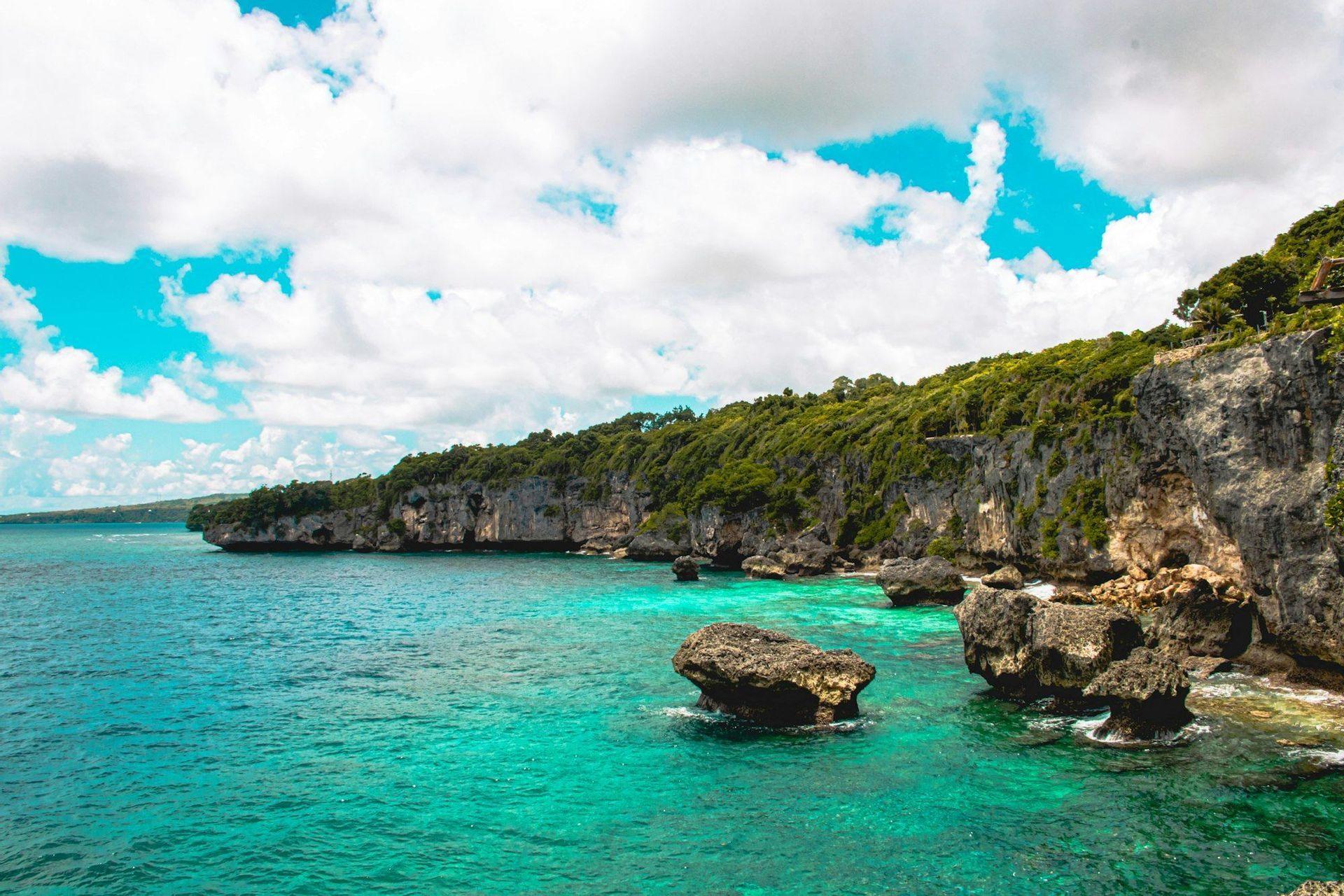 Une côte rocheuse couverte d'arbres verts longe une mer calme et turquoise avec de gros rochers sous un ciel partiellement nuageux.