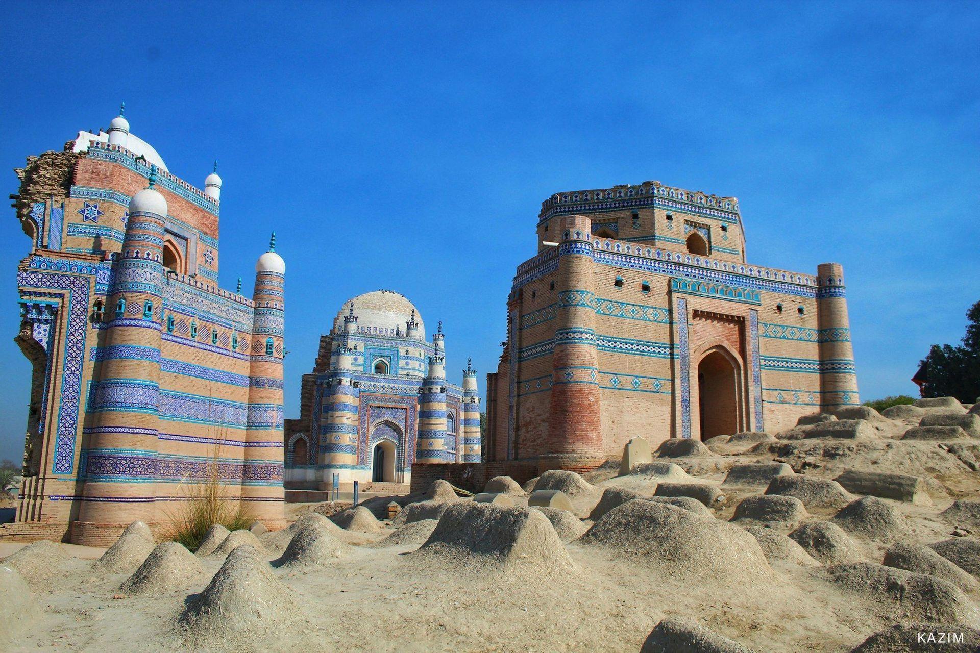 D'anciens mausolées décorés de carreaux bleus et ocre se dressent au sommet d'une colline recouverte de tombes en terre, sous un ciel d'un bleu éclatant.