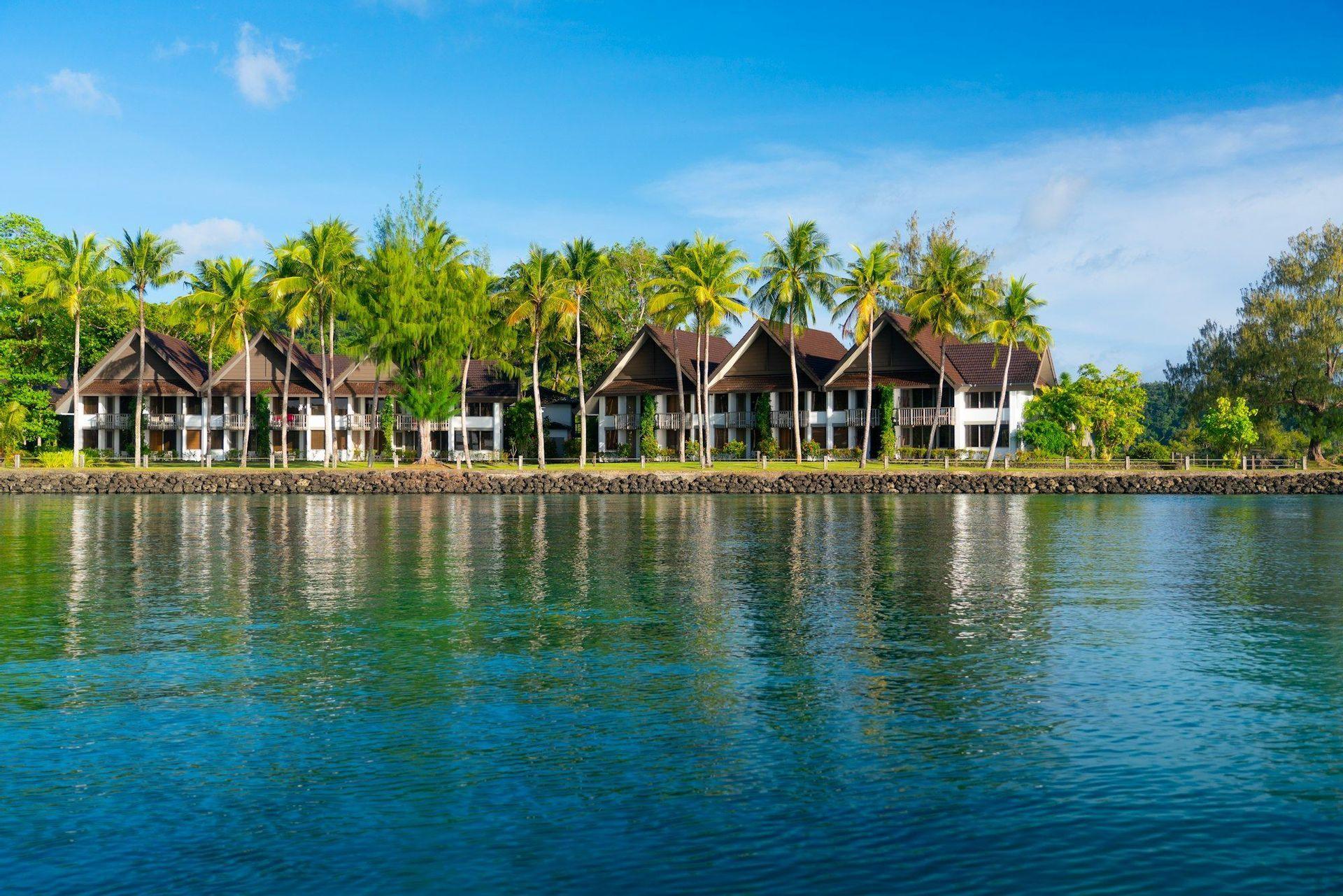 Une rangée de villas en bord de mer nichées parmi de grands palmiers, leurs reflets visibles sur l'eau bleue calme.