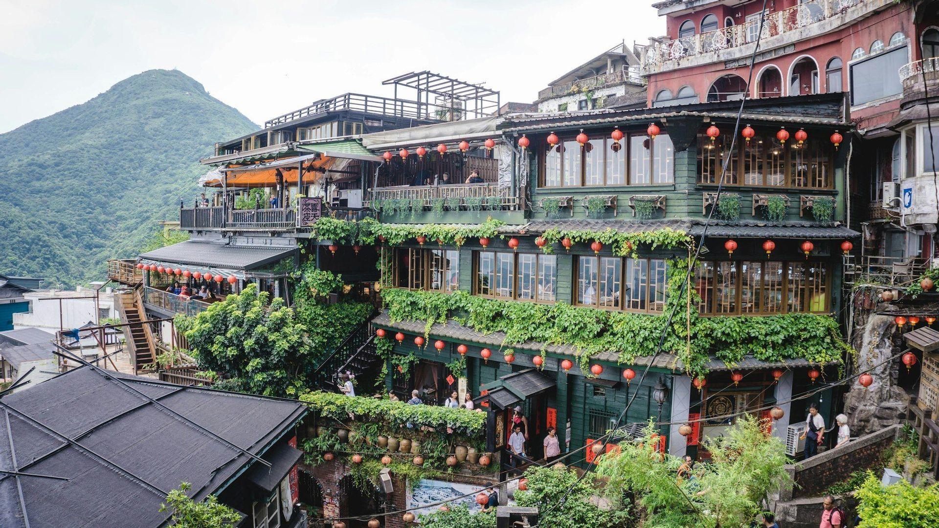 Un bâtiment vert élaboré à plusieurs étages, couvert de lierre et décoré de lanternes rouges, se détachant sur une montagne boisée.