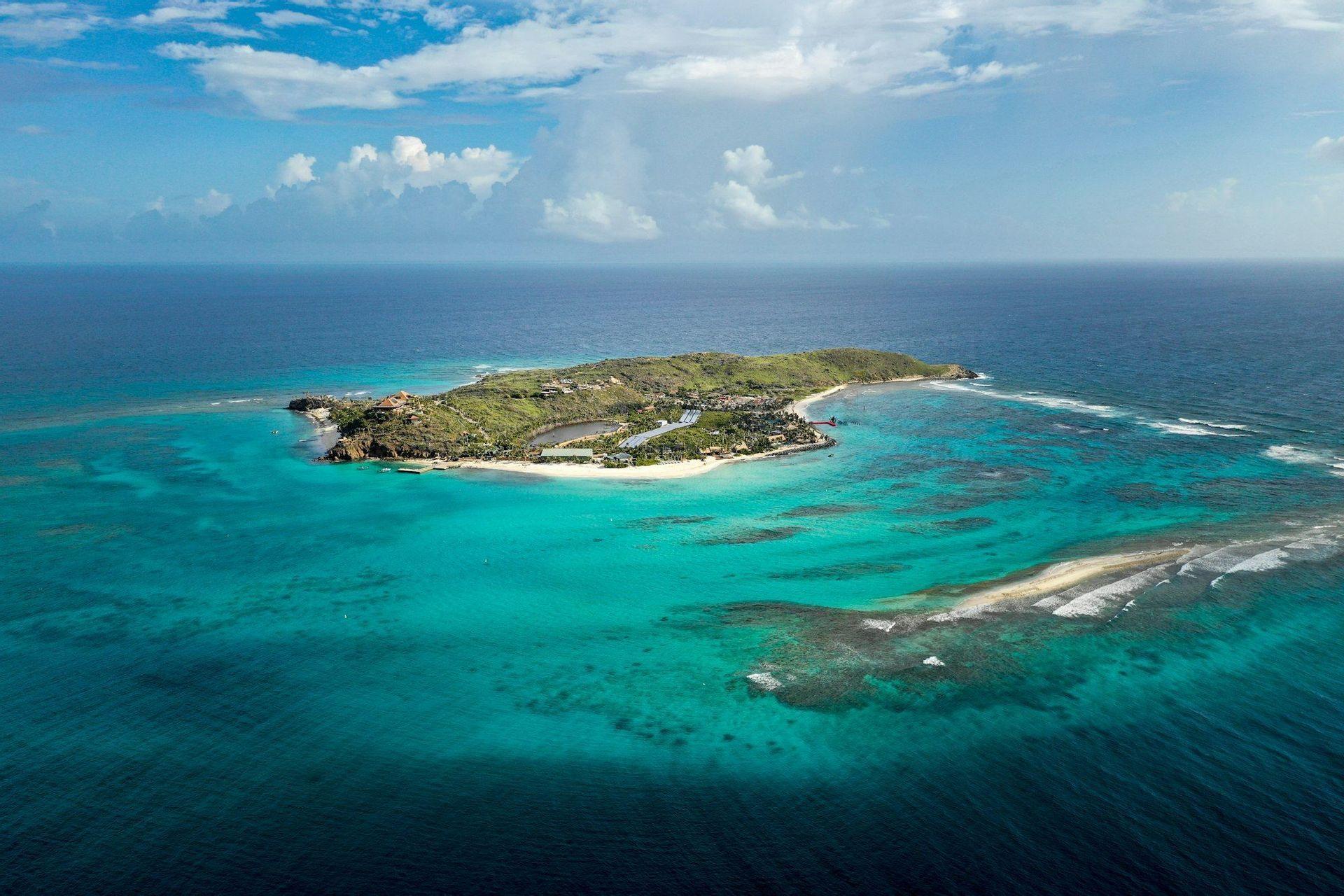 Vue aérienne d'une île verdoyante avec une plage de sable blanc, entourée d'eaux turquoise et de récifs coralliens sous un ciel partiellement nuageux.