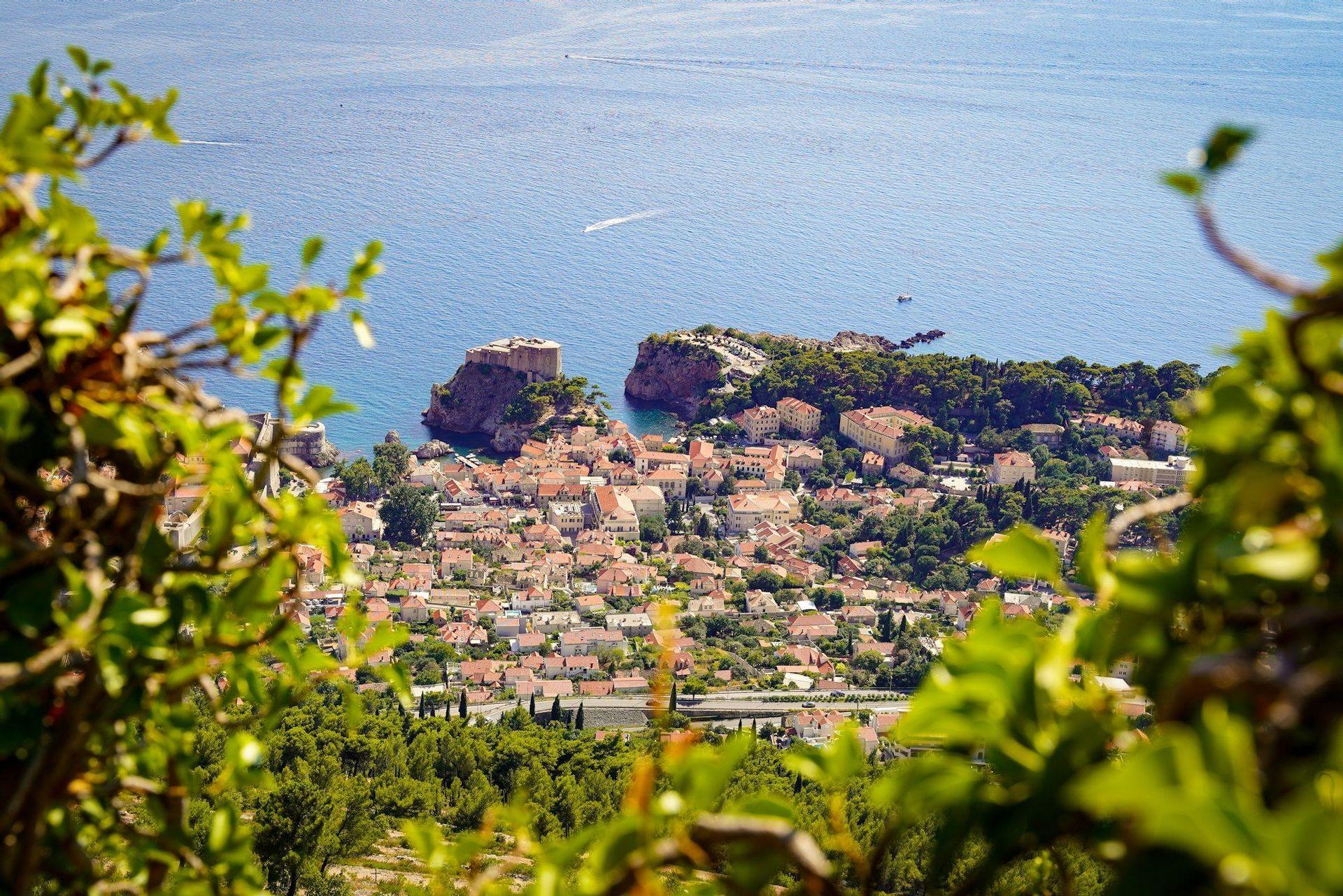 Une vue aérienne d'une ville côtière avec des bâtiments aux toits rouges et une forteresse sur une péninsule, encadrée par des feuilles vertes.