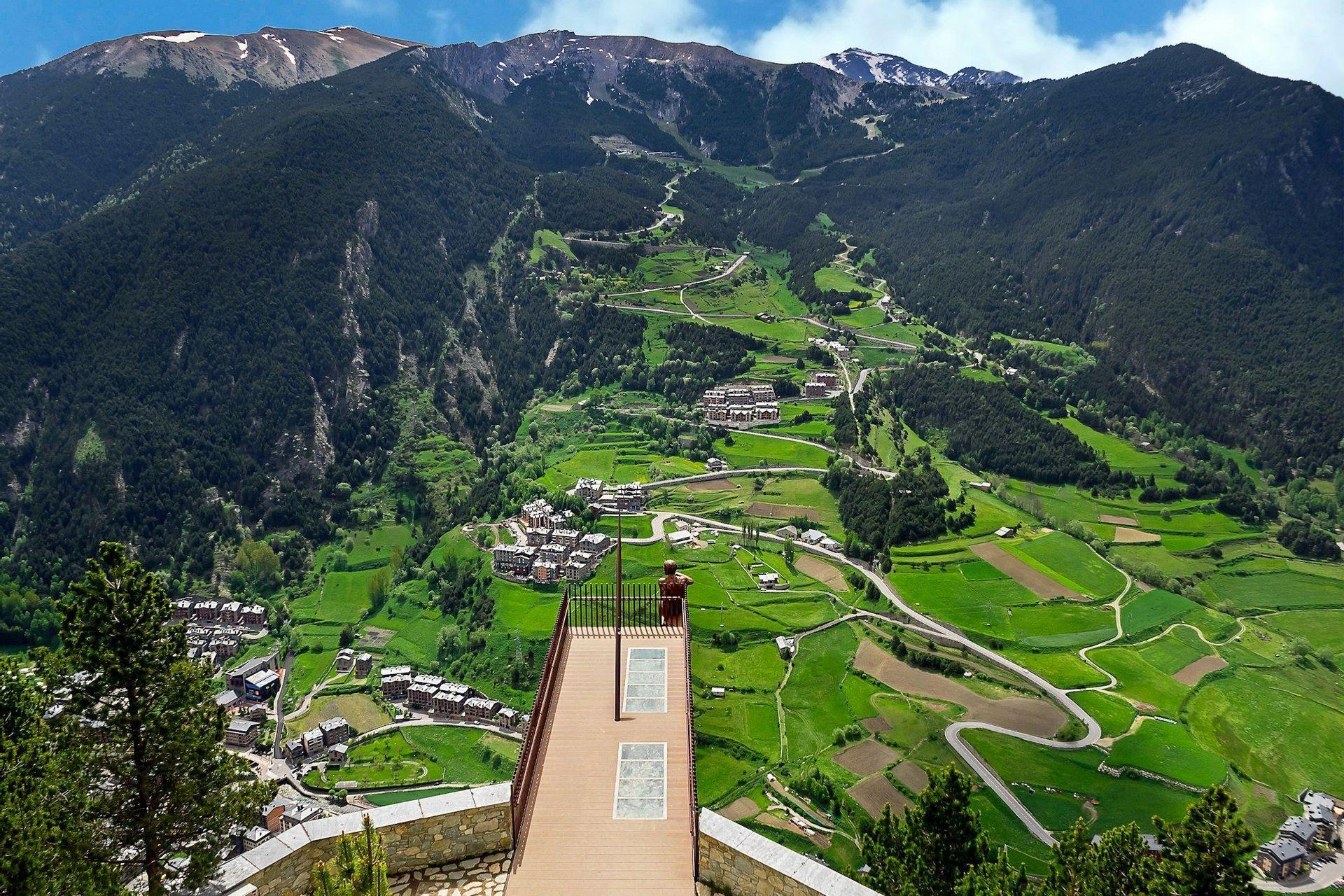 A person on a wooden viewpoint overlooks a vast green valley with villages, winding roads, and snow-capped mountains in the distance.