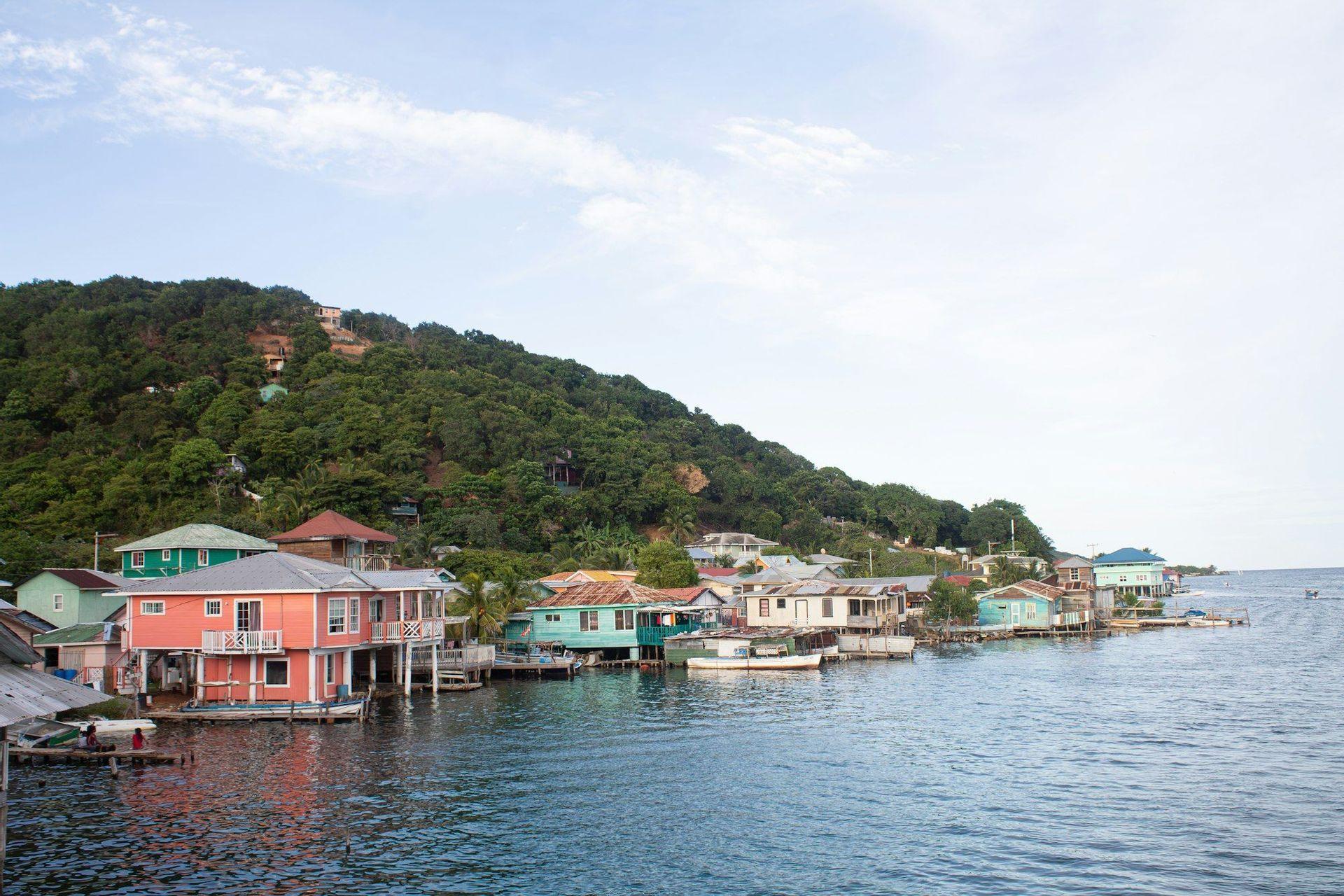 Colorful houses on stilts line the waterfront at the base of a lush, tree-covered hill under a bright sky.