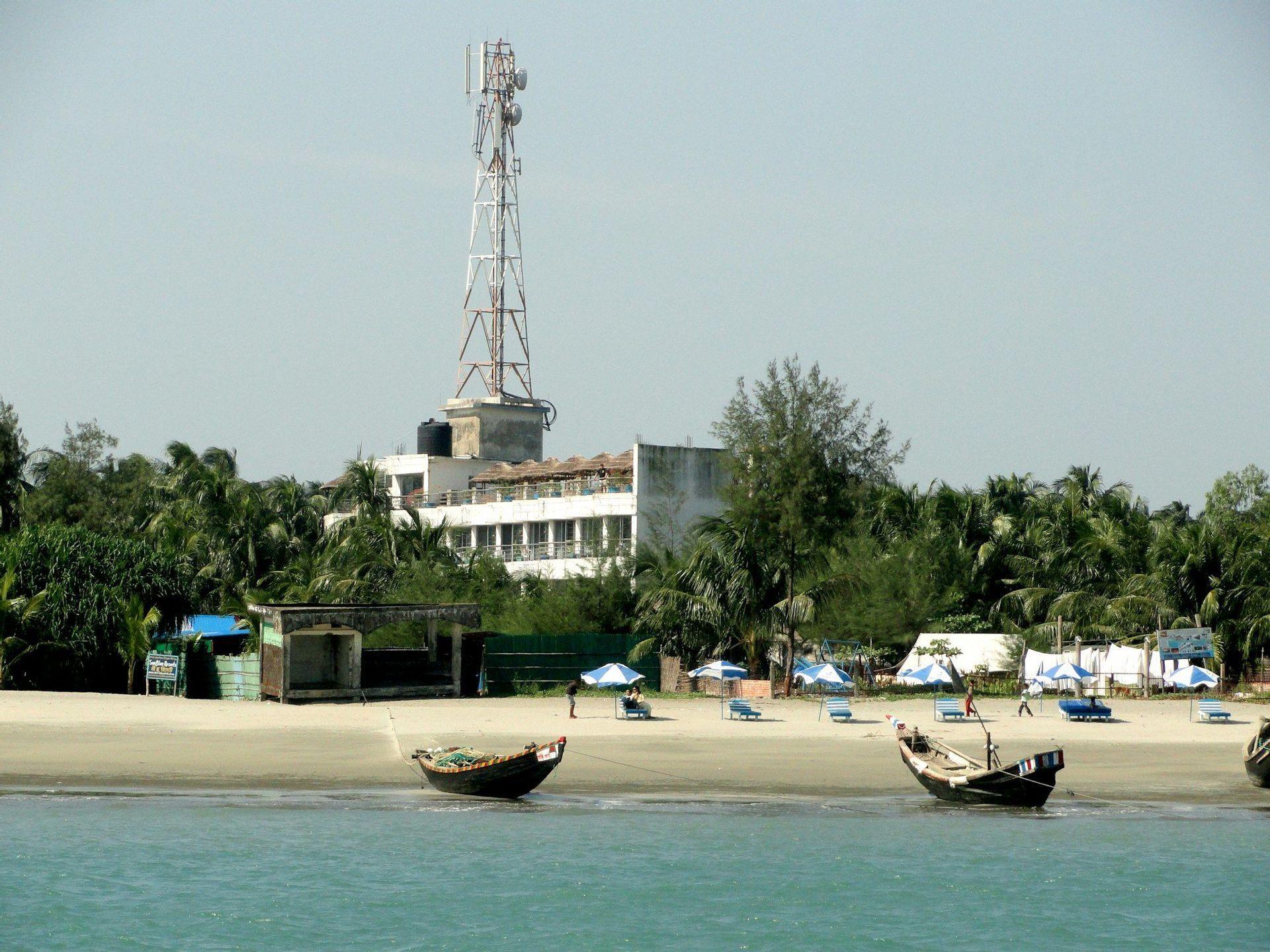 Two wooden boats rest in the shallows of a sandy beach lined with palm trees, with a building and a tall communications tower in the background.
