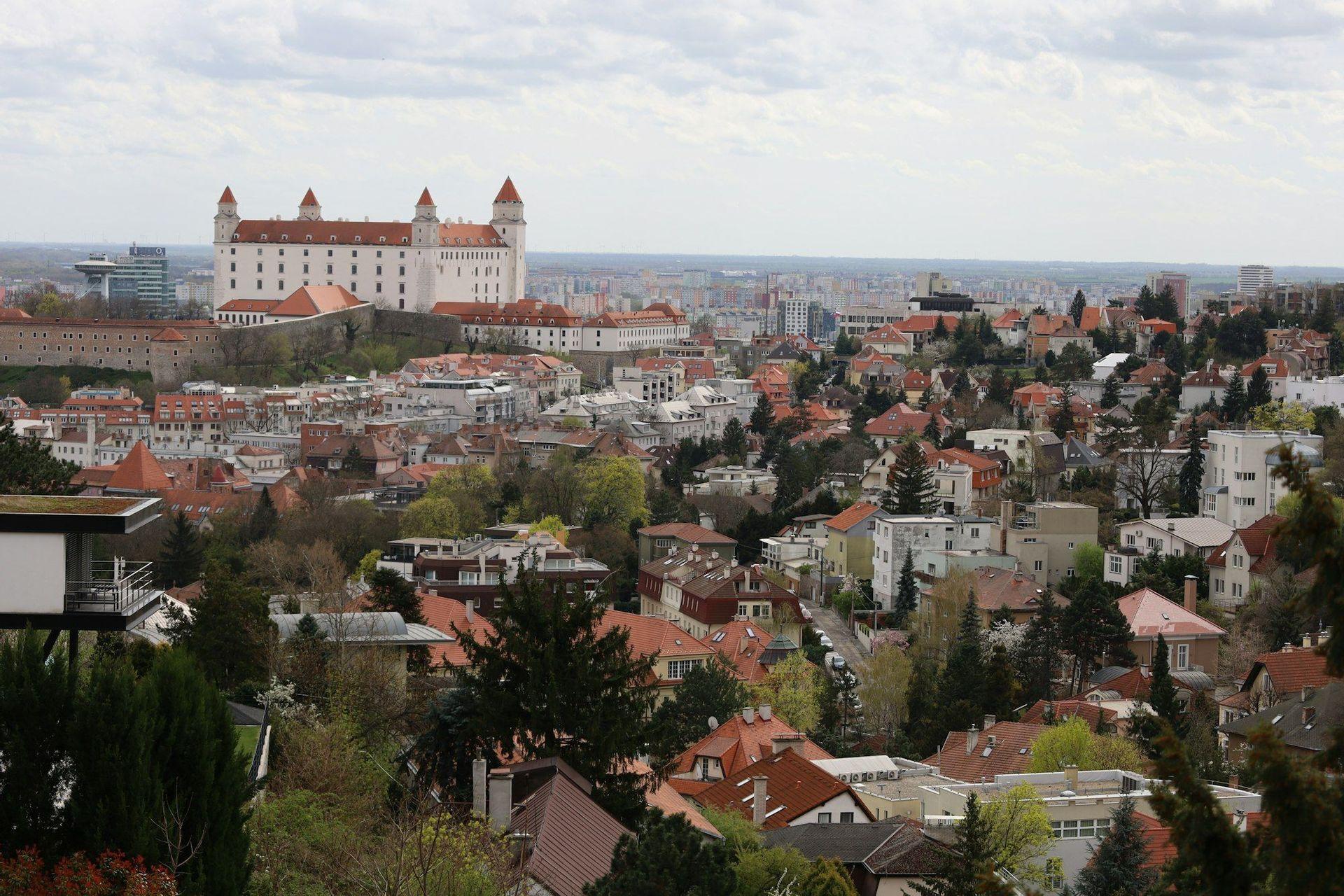 A large, white castle with red-roofed towers sits on a hill above a sprawling cityscape with trees under a cloudy sky.