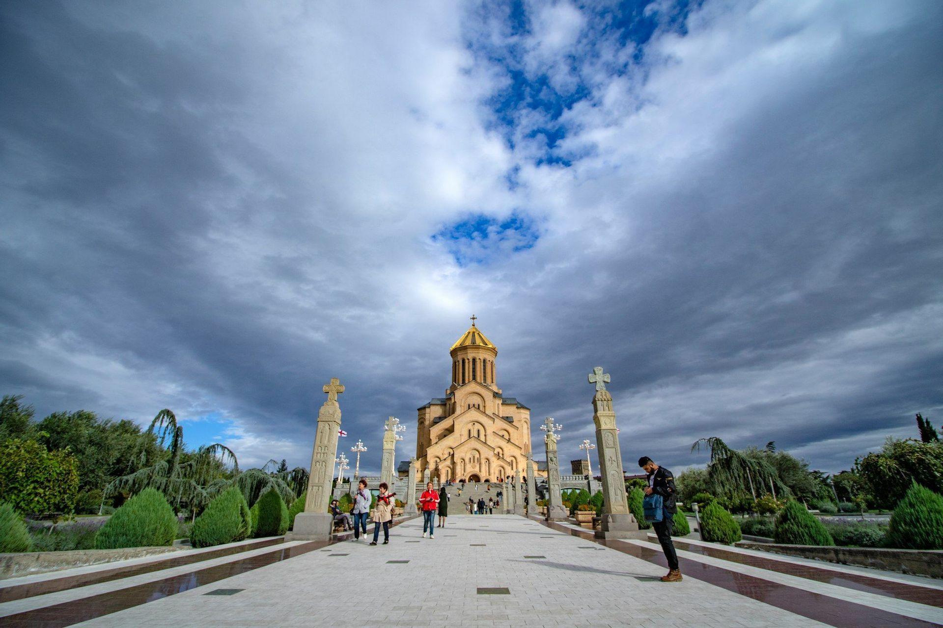 A wide-angle view of a stone cathedral with a golden dome, with people walking along a path lined with crosses under a dramatic cloudy sky.