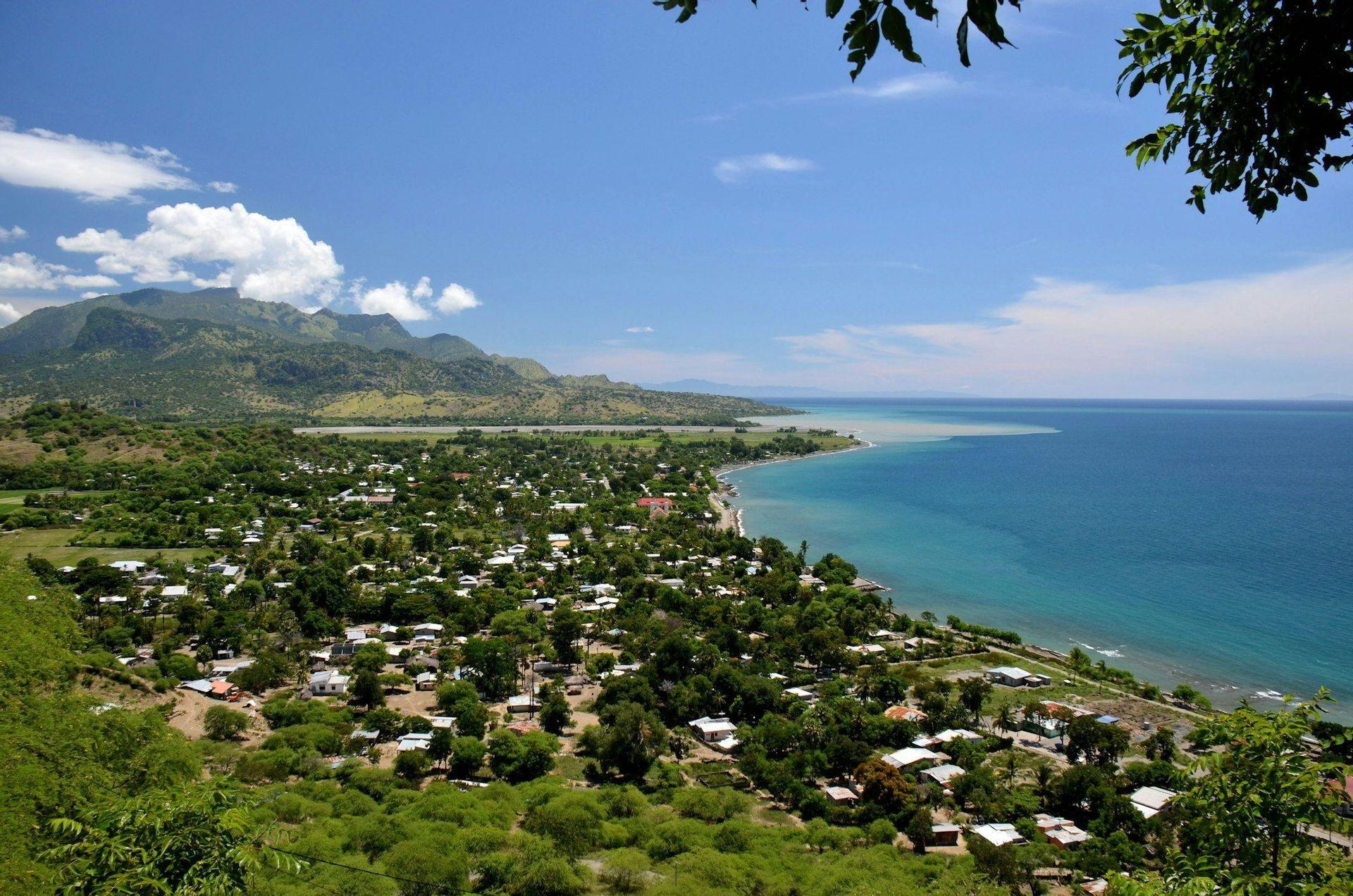 An aerial view of a coastal village surrounded by green trees, with large mountains in the background and a calm blue ocean.