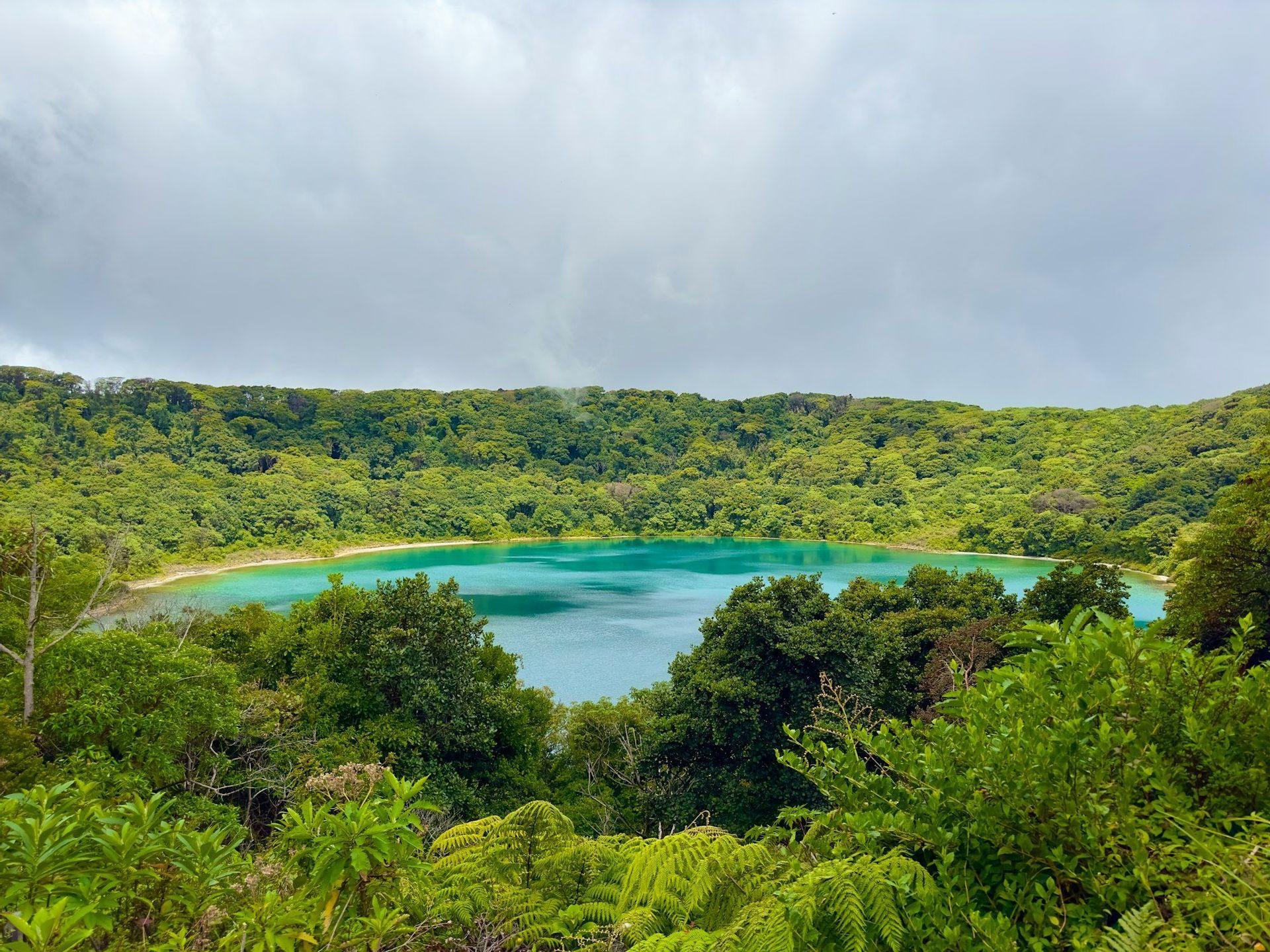 A high-angle view of a turquoise crater lake surrounded by dense, green forest under a cloudy sky.