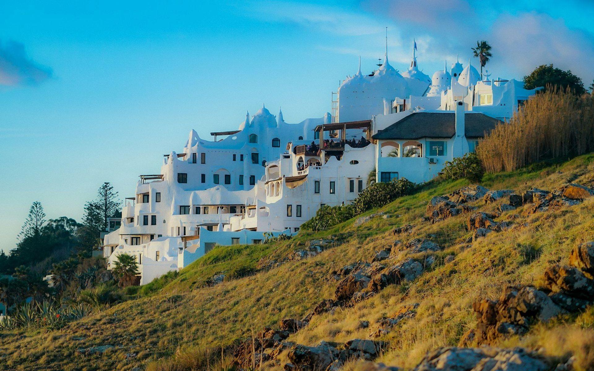 A unique, whitewashed building complex with sculpted features sits on a grassy, rocky hillside under a clear blue sky.