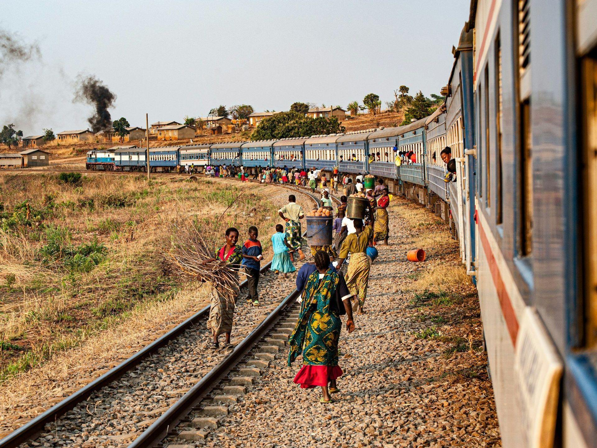 A crowd of people walk along the tracks beside a long blue train, with some carrying goods on their heads and in baskets.