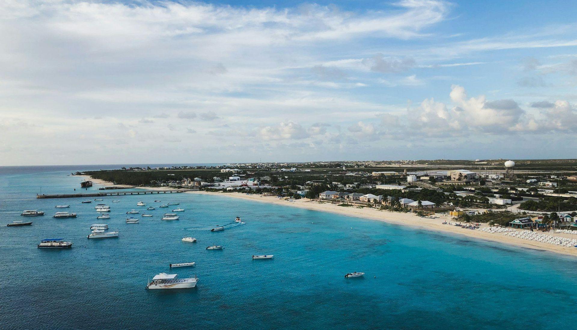 An aerial view of a coastal town with numerous boats floating in the turquoise sea alongside a sandy beach.