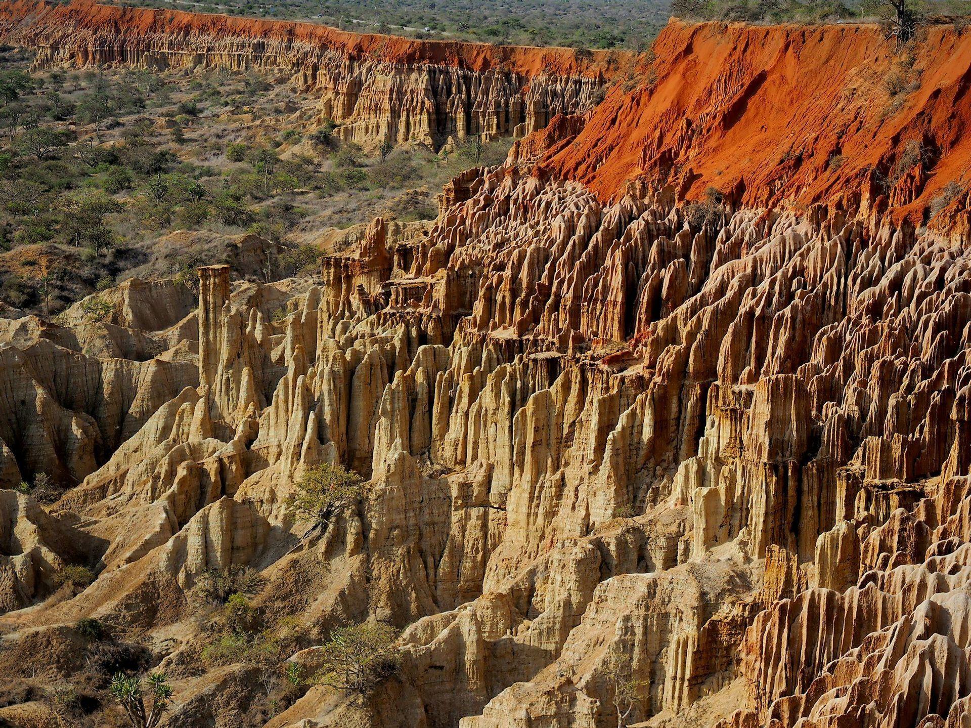 A landscape of beige and tan eroded rock pinnacles and spires, with a bright red earth layer on the upper ridges.