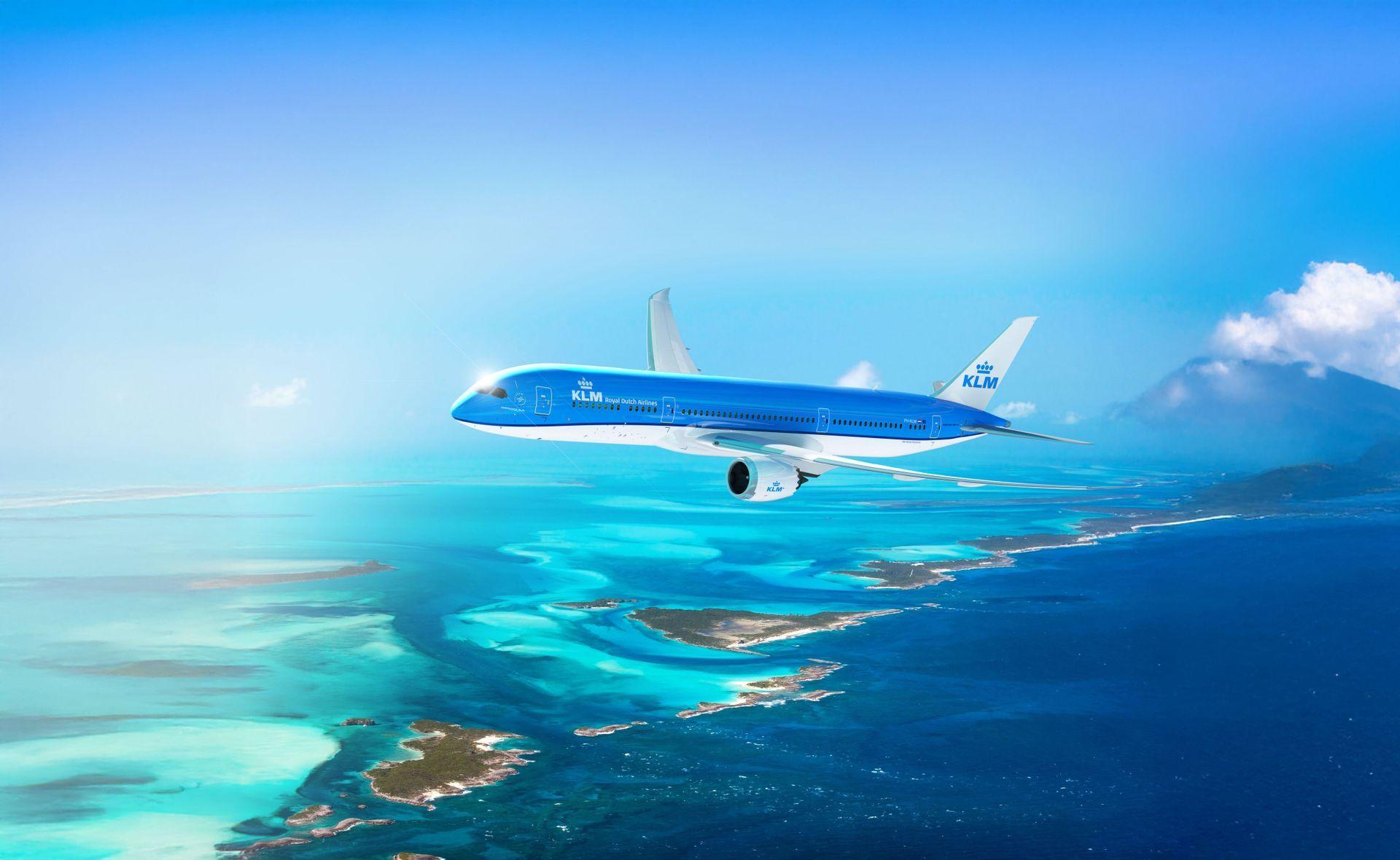 Un aereo blu della KLM vola sopra un oceano turchese disseminato di piccole isole verdi sotto un cielo azzurro brillante.