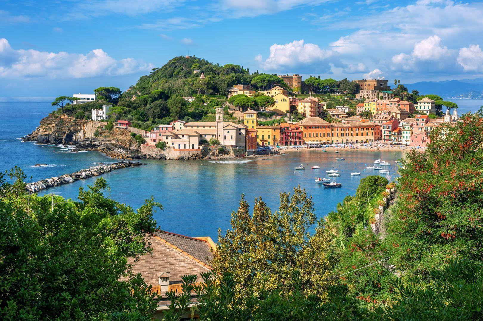 Colorful buildings line a seaside town's bay filled with boats, set against a green peninsula under a blue sky with clouds.