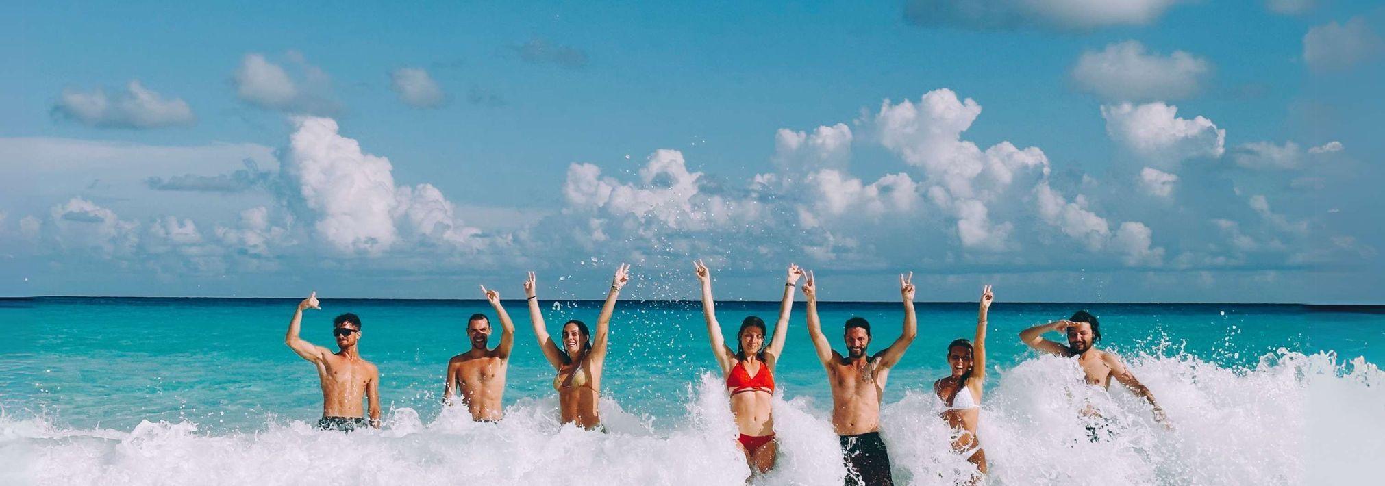 Un grupo de WeRoad de pie en las olas turquesas del mar, sonriendo con los brazos en alto y haciendo el signo de la paz.