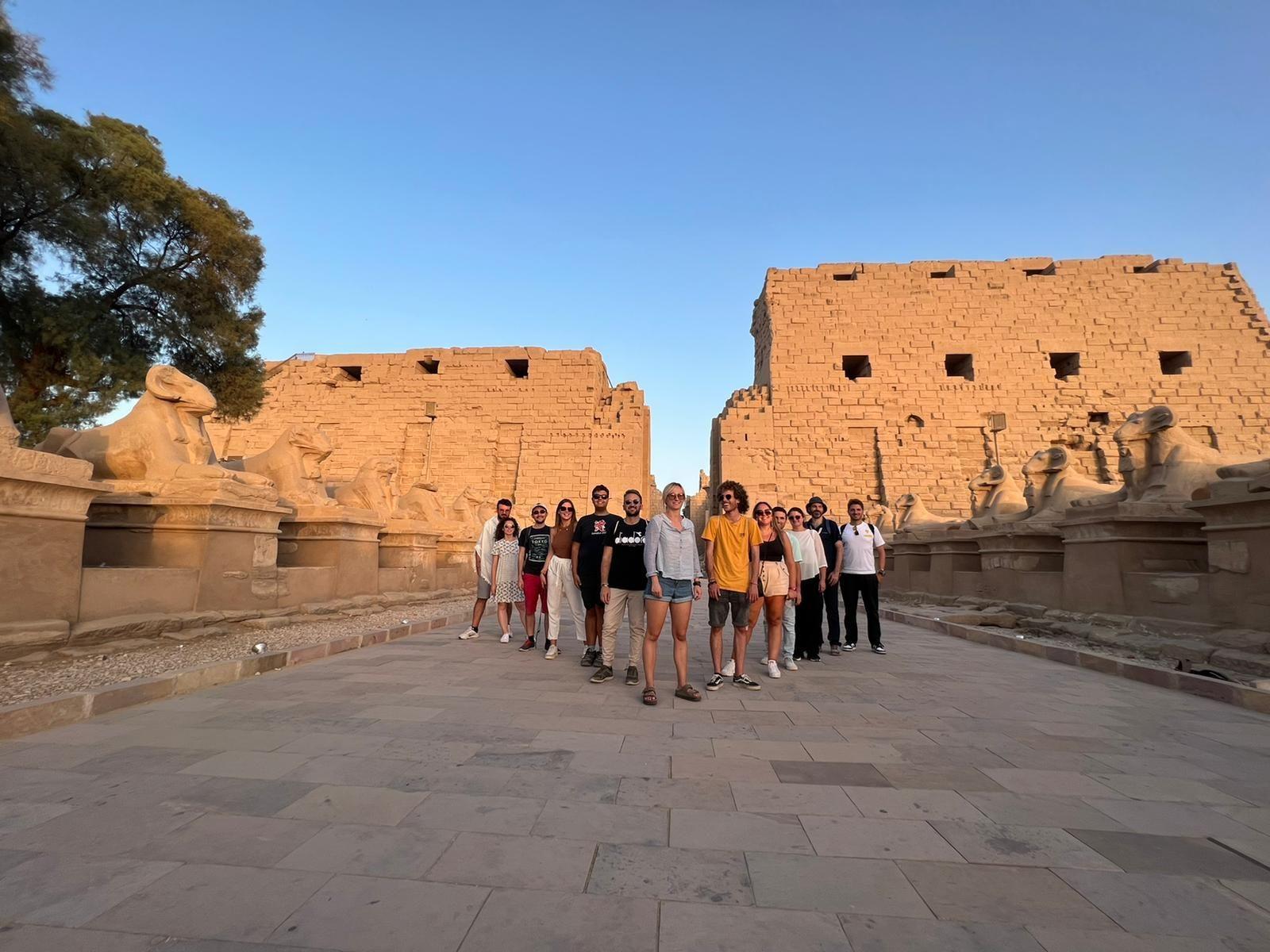 Un gruppo WeRoad in viaggio si trova su un viale di pietra tra due grandi piloni di un tempio, affiancato da statue di sfingi con teste di ariete.