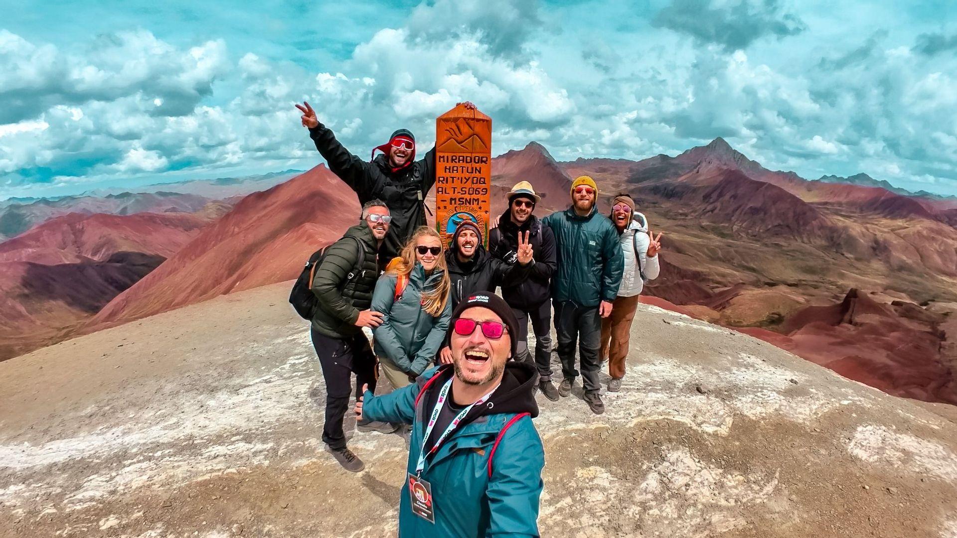 Un grupo de WeRoad se toma una selfie en la cima de una montaña con un panorama de coloridas montañas rojas de fondo.