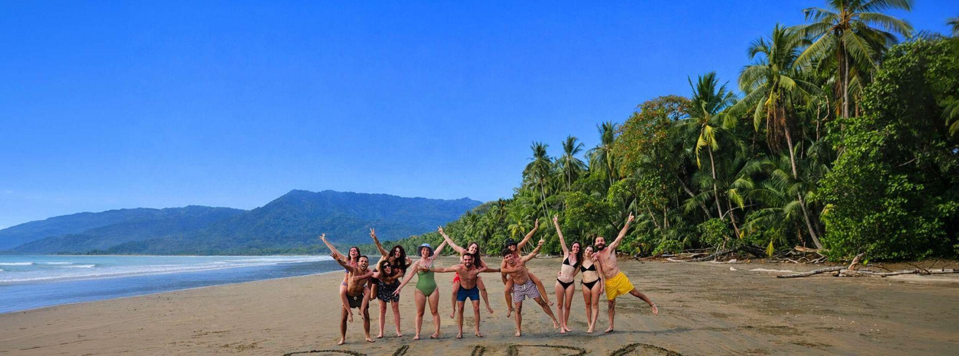 Un viaggio di gruppo WeRoad in posa insieme in costume da bagno su una spiaggia tropicale fiancheggiata da palme e una giungla.