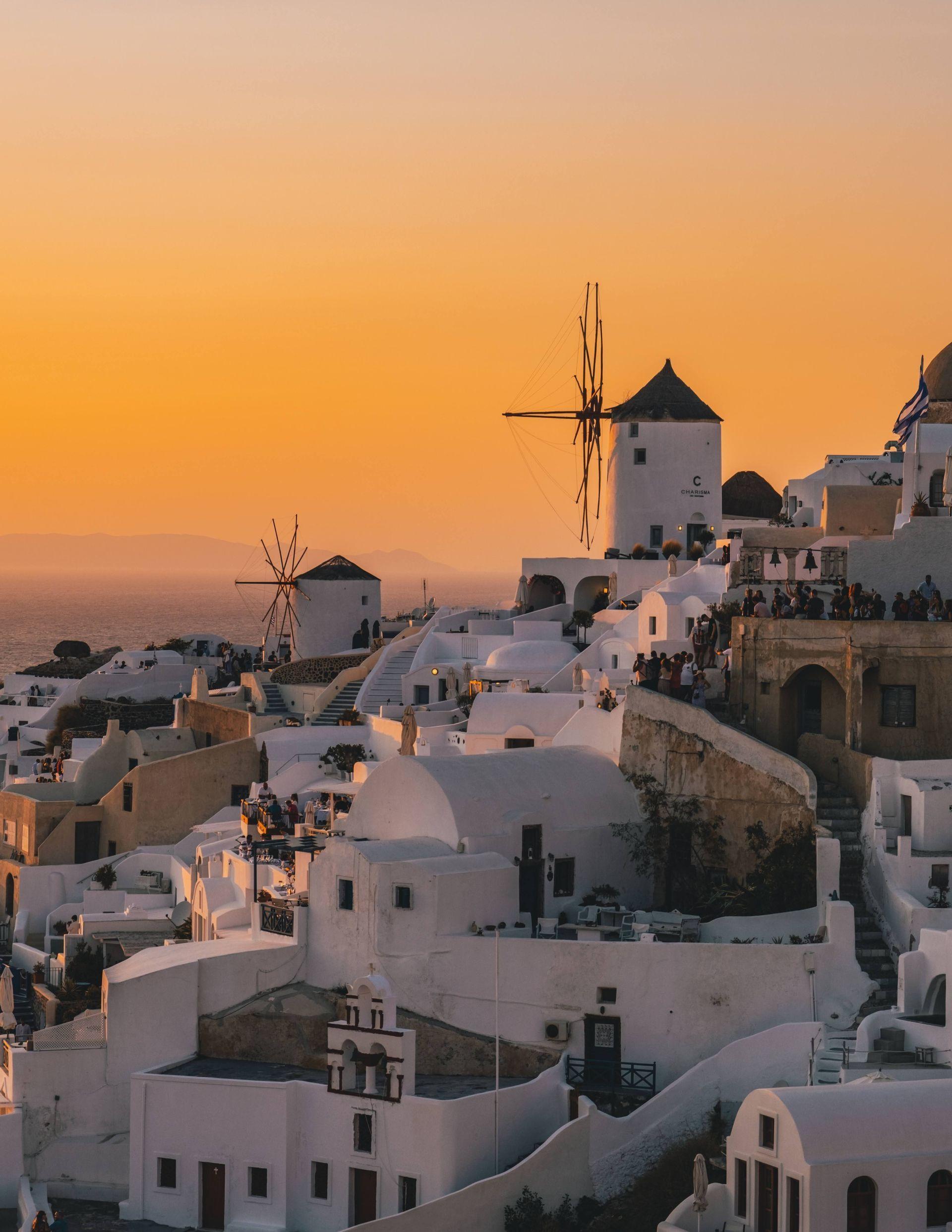 Edificios encalados y molinos de viento tradicionales se apiñan en un acantilado con vistas al mar bajo un cielo de atardecer dorado.