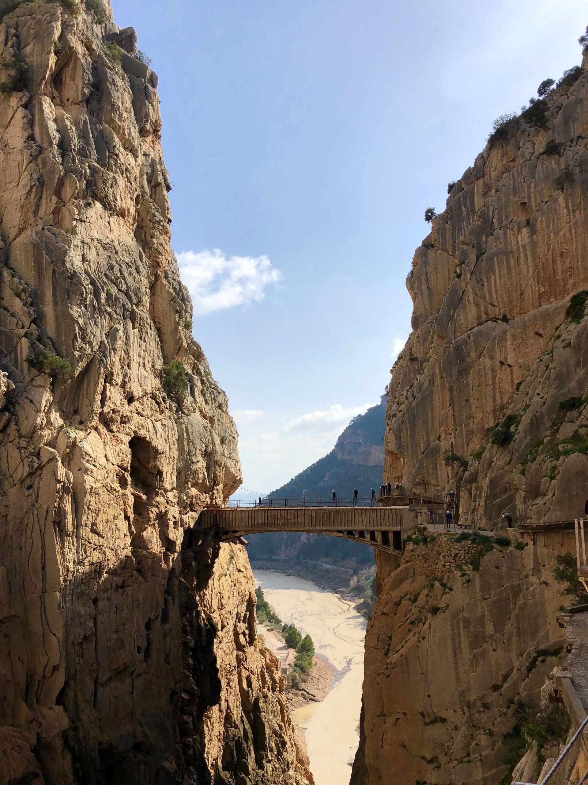 A WeRoad group trip walks across a narrow bridge suspended between two sheer rock walls of a canyon, with a river far below.