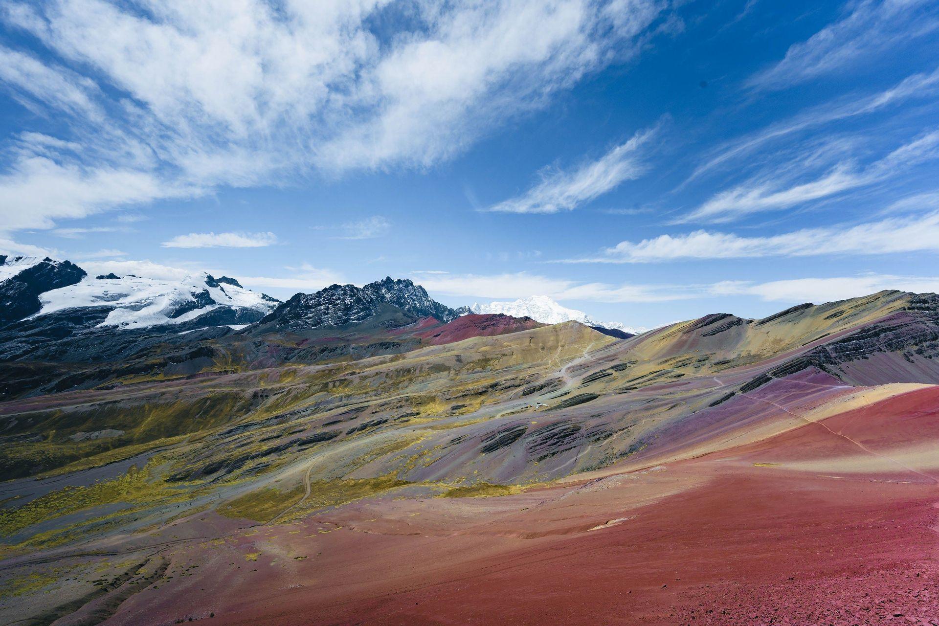 Un vaste paysage de montagnes colorées et rayées avec des sommets enneigés en arrière-plan, sous un ciel bleu et nuageux.