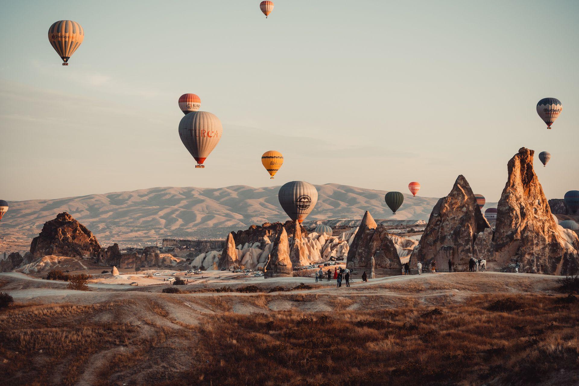 Heißluftballons schweben über einer Landschaft aus einzigartigen Felsformationen, während eine Gruppe von Menschen vom Boden aus zuschaut.