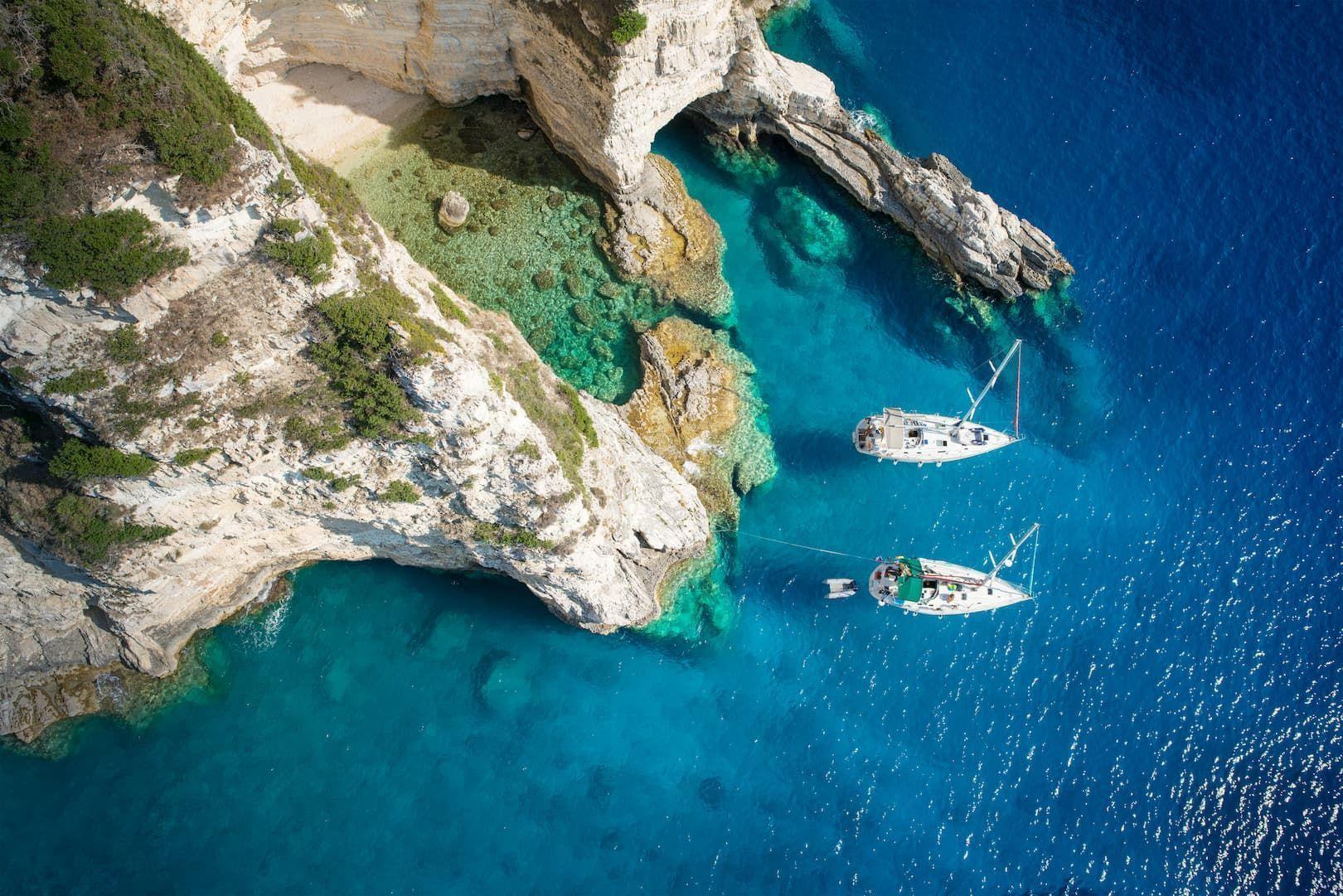 An aerial view of two sailboats anchored in turquoise water next to white rocky cliffs and a secluded cove.