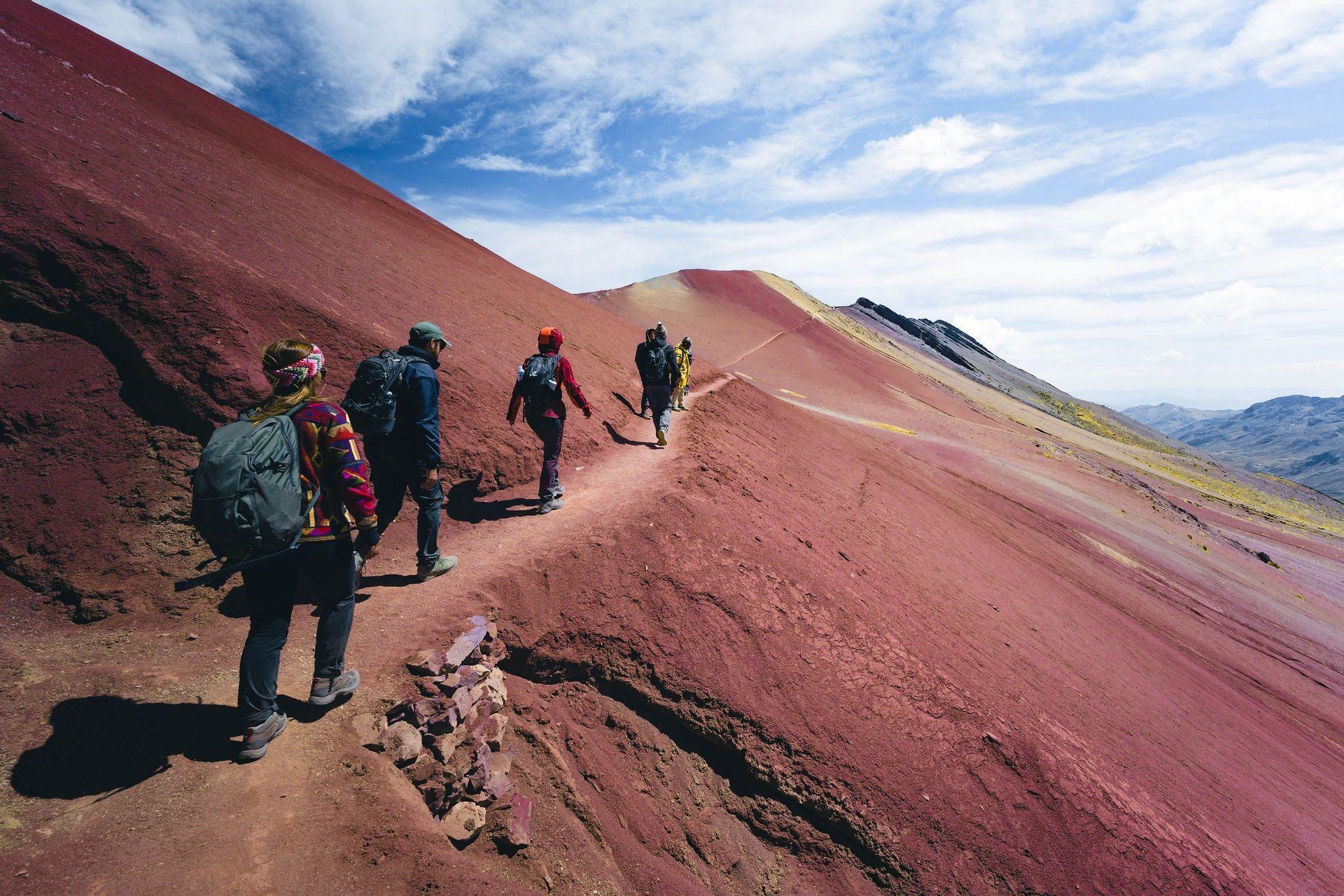 A WeRoad group trip hikes in single file along a narrow trail on a steep, red mountain under a blue sky.