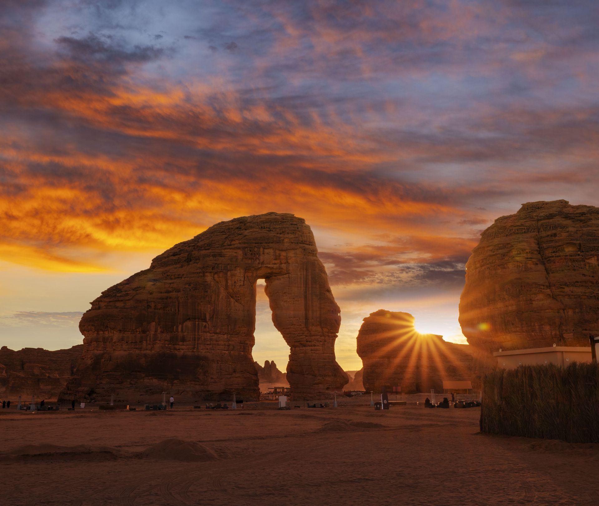 Ein großer natürlicher Felsbogen in einer Wüstenlandschaft bei Sonnenuntergang, wobei die Sonne einen Strahlenkranz-Effekt vor einem farbenfrohen Himmel erzeugt.