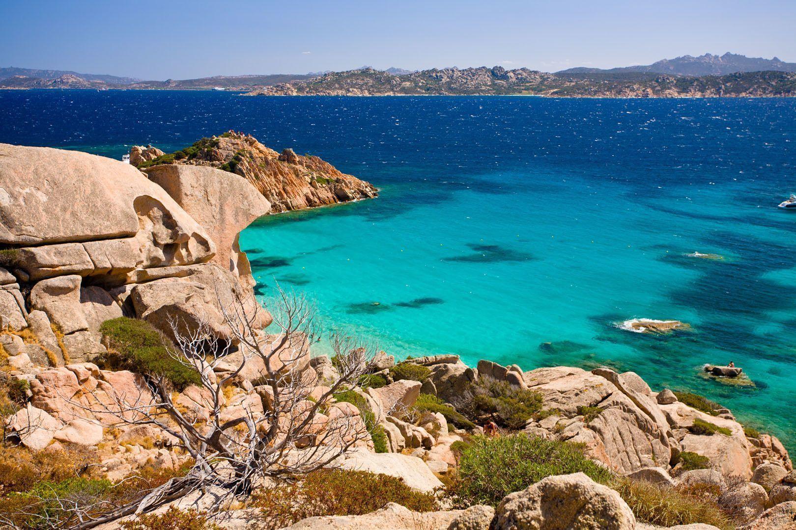 A scenic view from a rocky coast overlooking a bay with clear turquoise water, with the deep blue sea and a distant shoreline in the background.