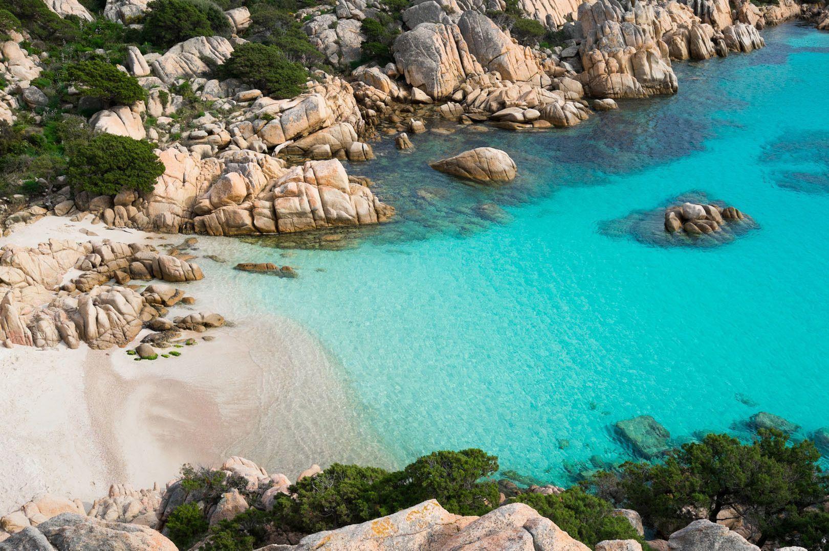 A secluded sandy cove with clear turquoise water is surrounded by large rocks and green shrubs, viewed from above.