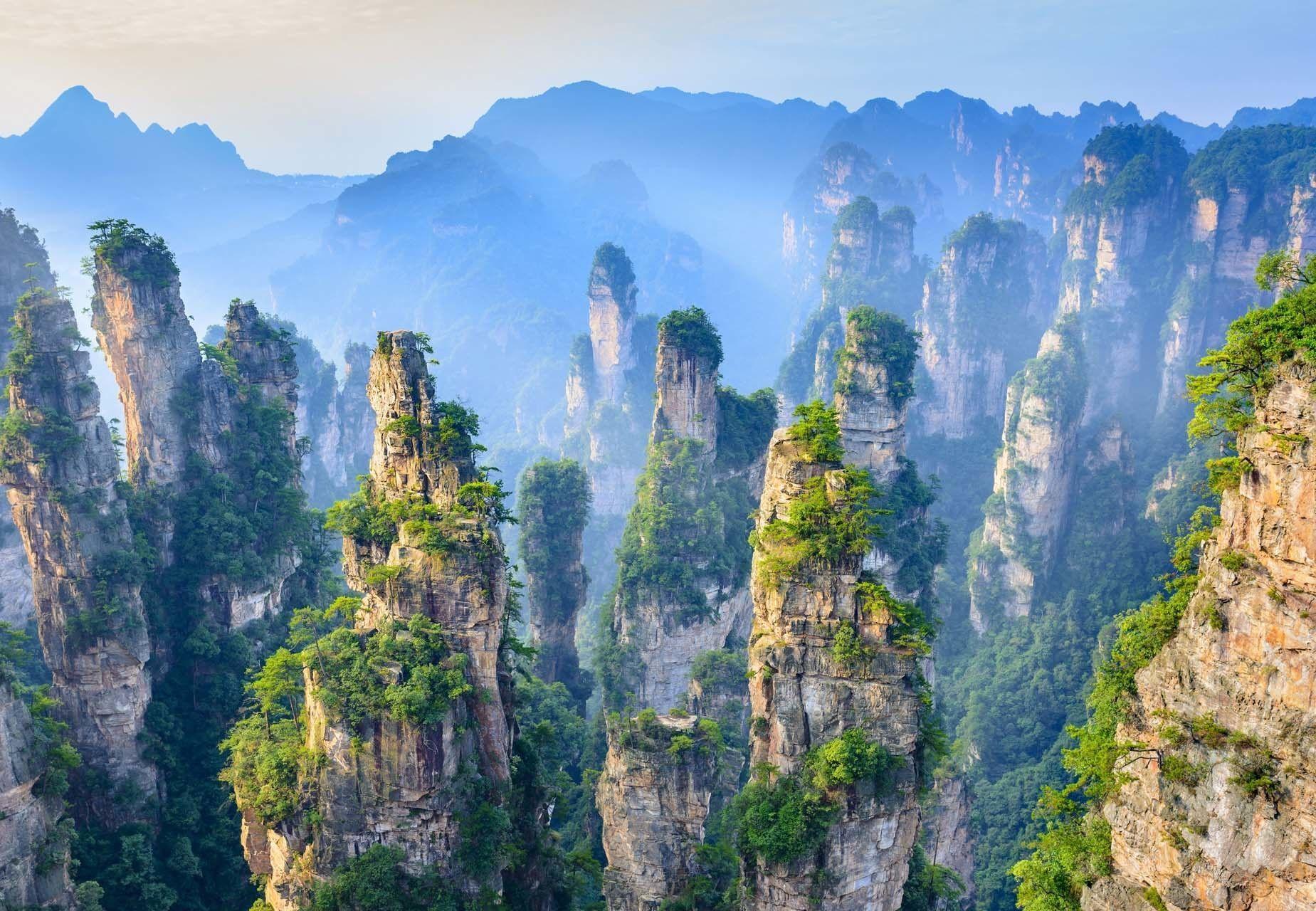Tall, pillar-like rock formations covered in green trees rise from a lush valley under a hazy blue sky.