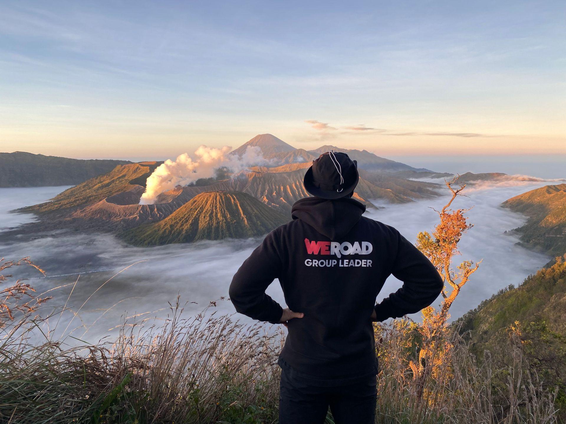 Una persona con una sudadera con capucha de WeRoad Group Leader observa el amanecer sobre un volcán humeante y un valle lleno de nubes.